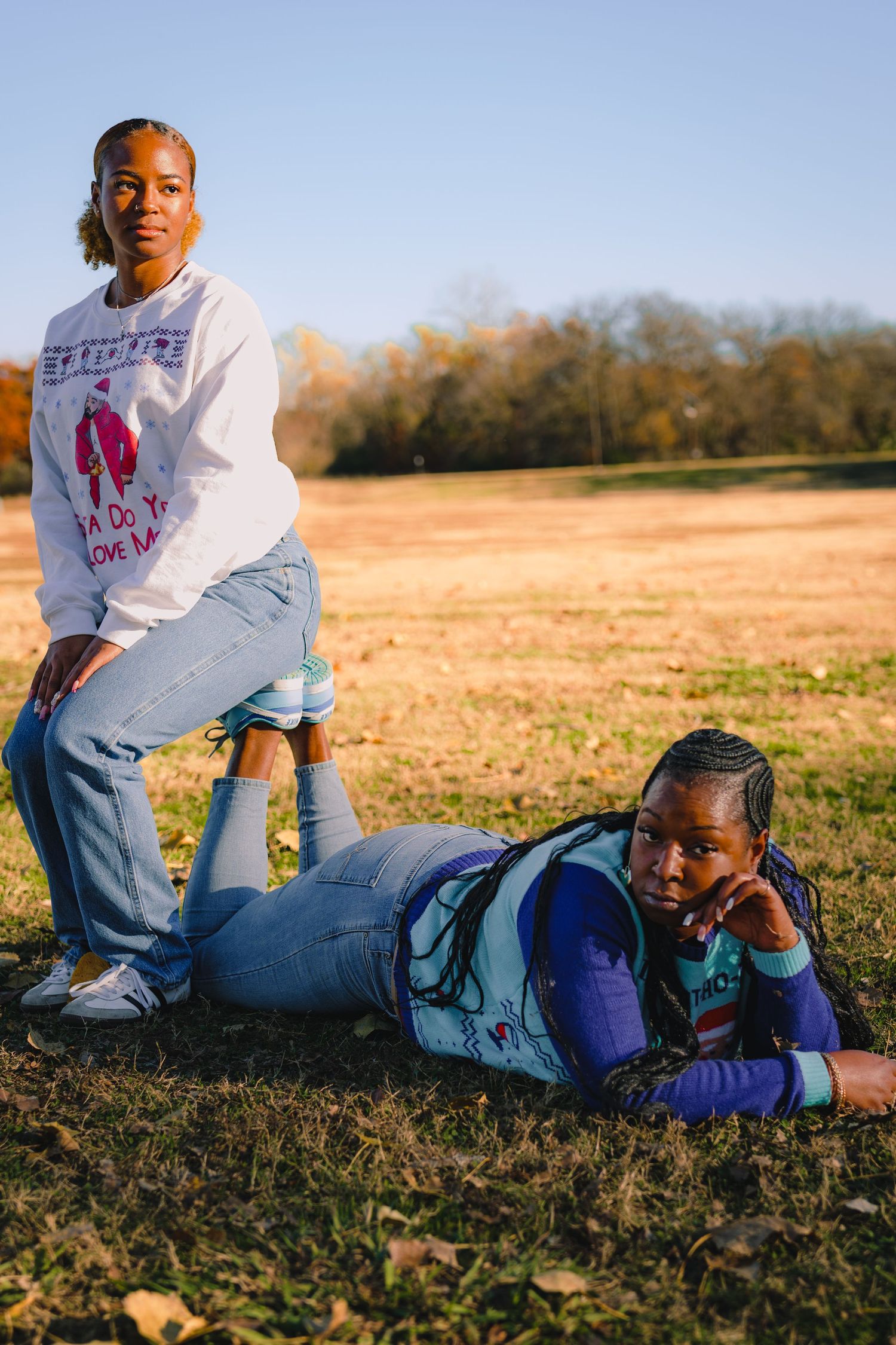 Two people in matching holiday sweaters pose casually on grass during sunset.