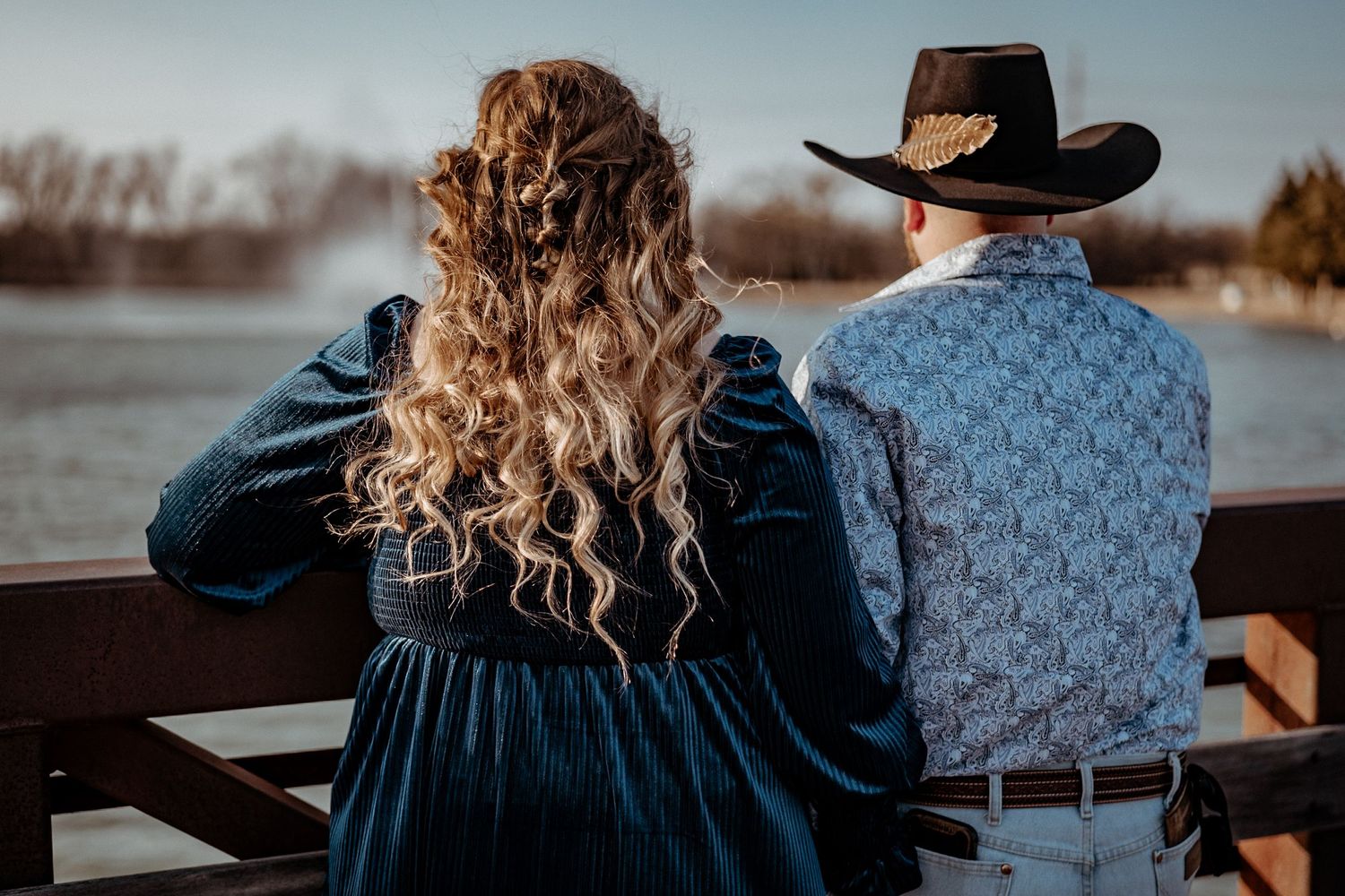 Couple in western attire sitting on bench overlooking water at sunset.