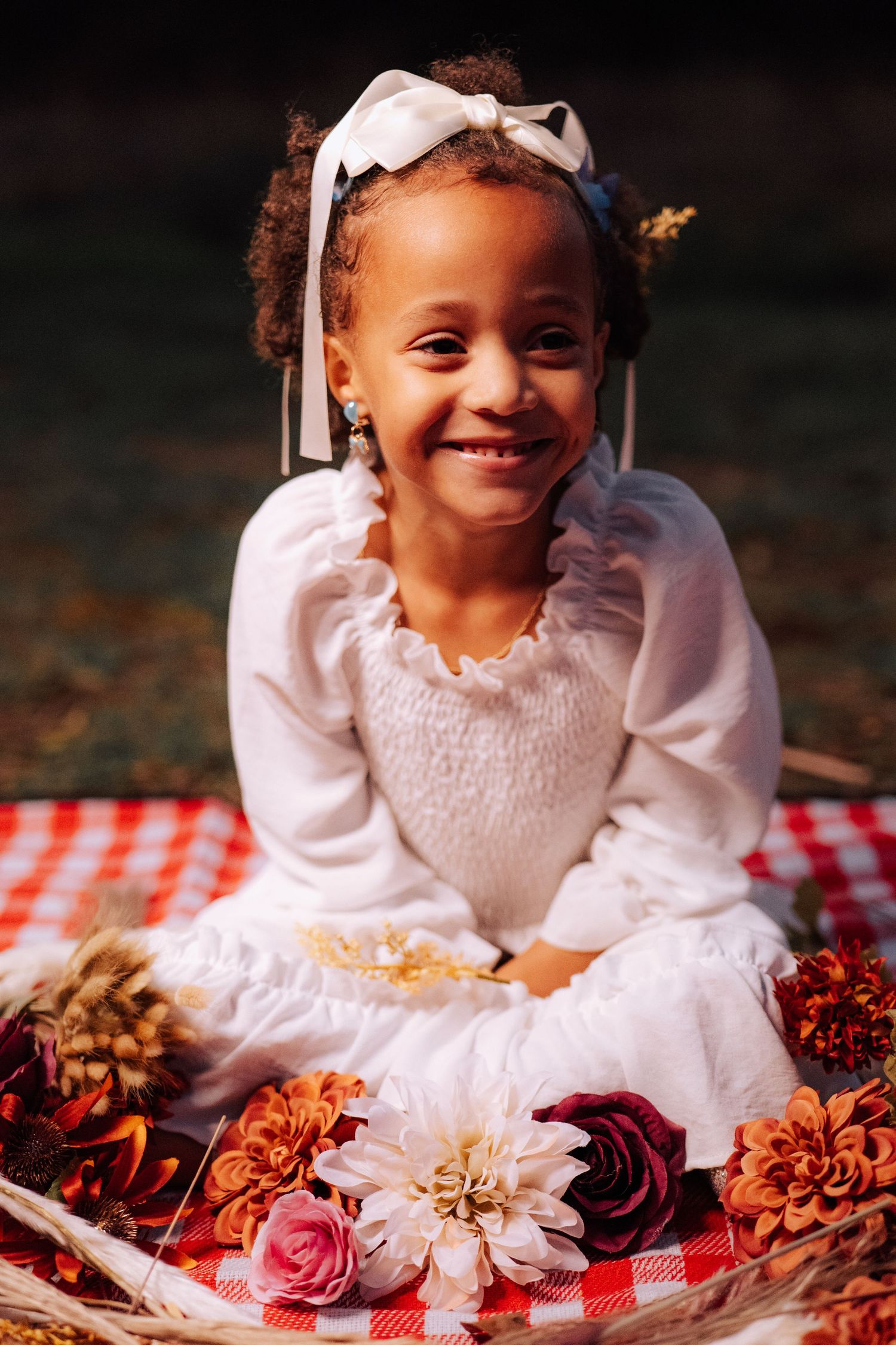 Portrait of a small child in white dress sitting on red checkered blanket surrounded by flowers.