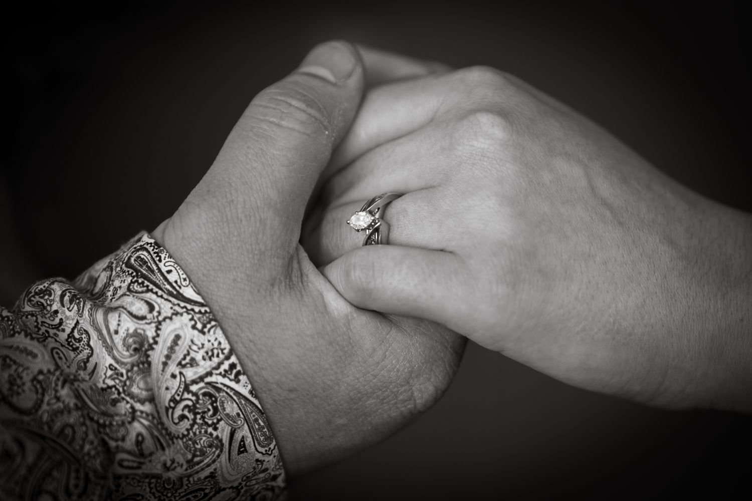 A close-up black and white photo showing entwined hands with one displaying intricate henna designs and a ring.