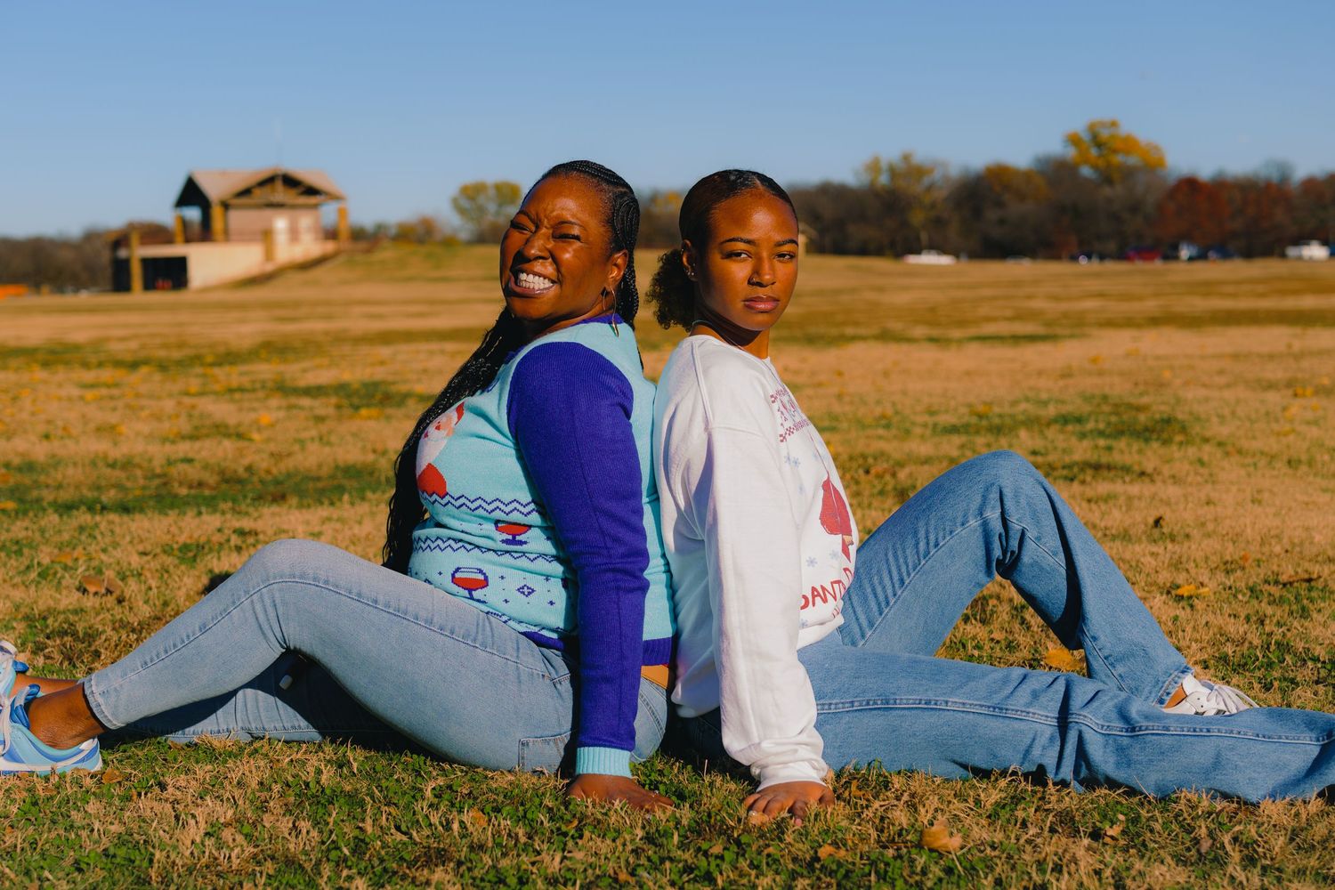 Two people in casual wear sitting back-to-back in a sunny field with a building in background.