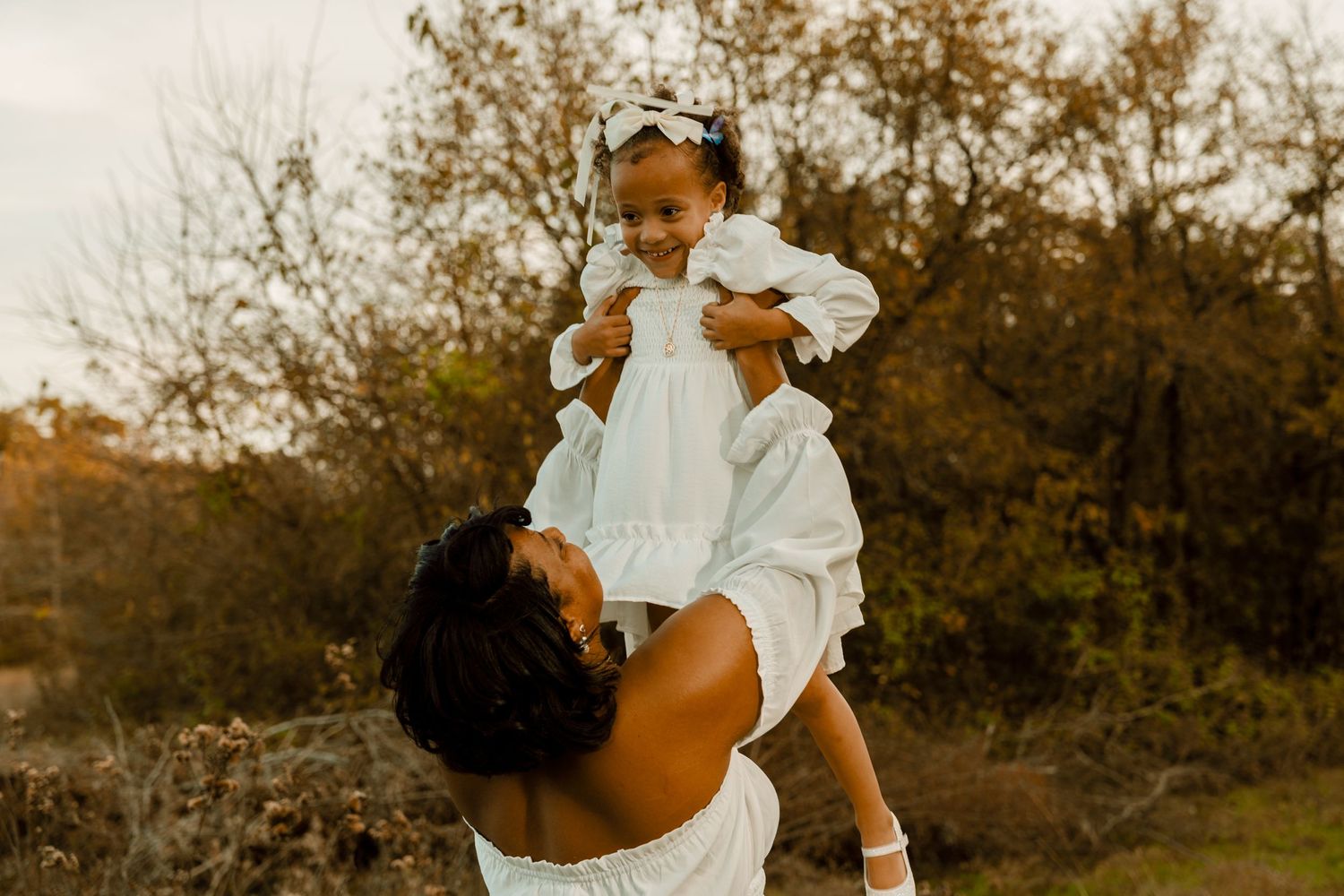 Mother in white dress lifting laughing child during outdoor autumn photoshoot.