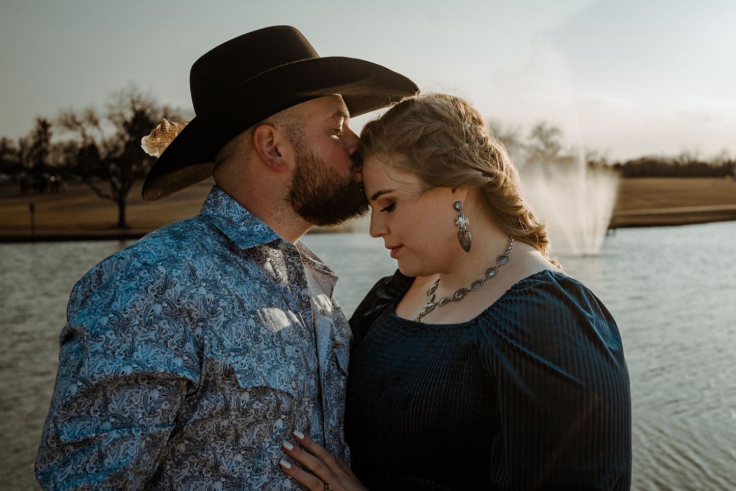 A tender moment between a couple in western wear by a lake at sunset.