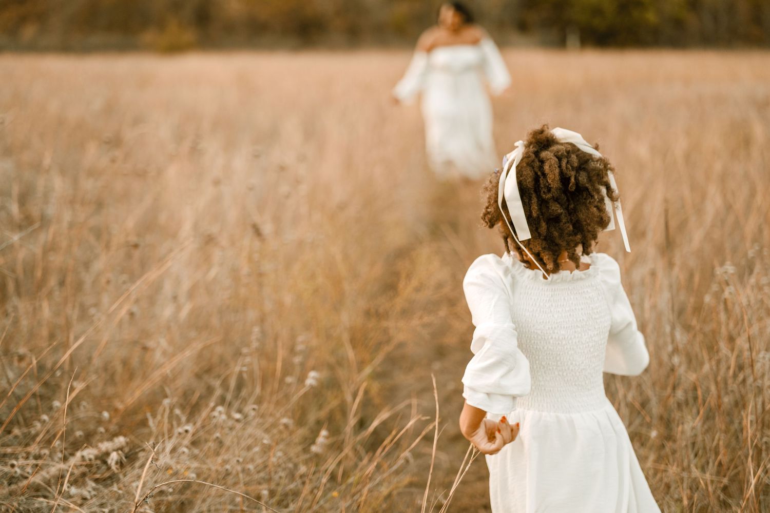 A dreamy back view of two figures in white dresses walking through tall golden grass.