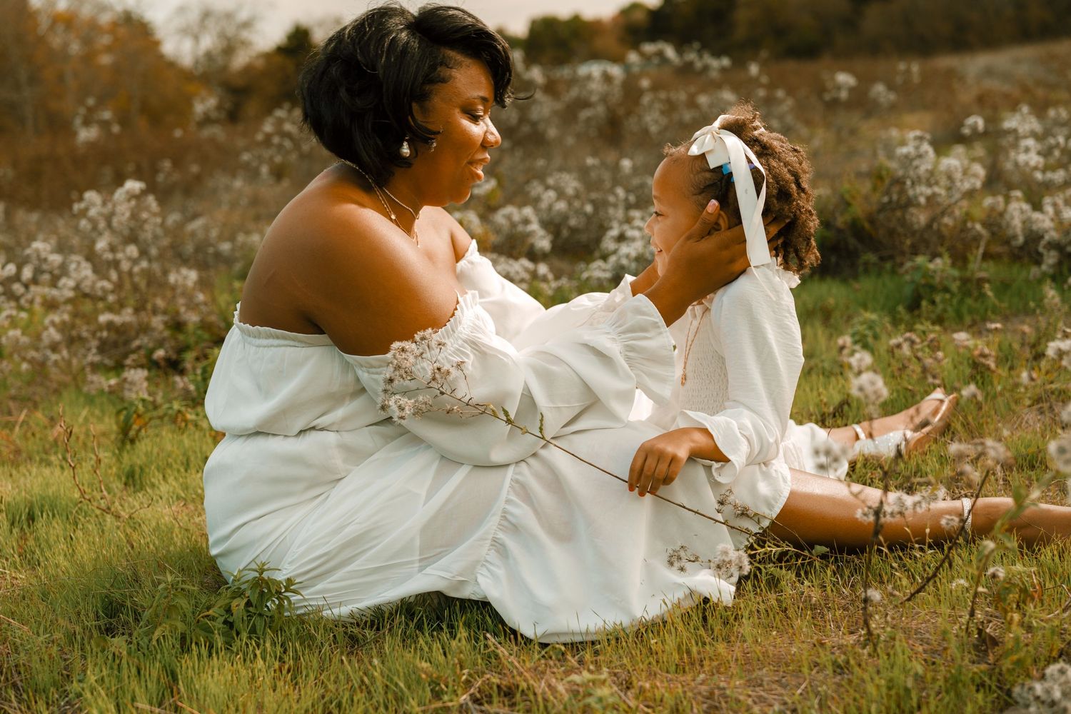 Two people in matching white dresses sit together on grassy ground during a warm sunset photoshoot.