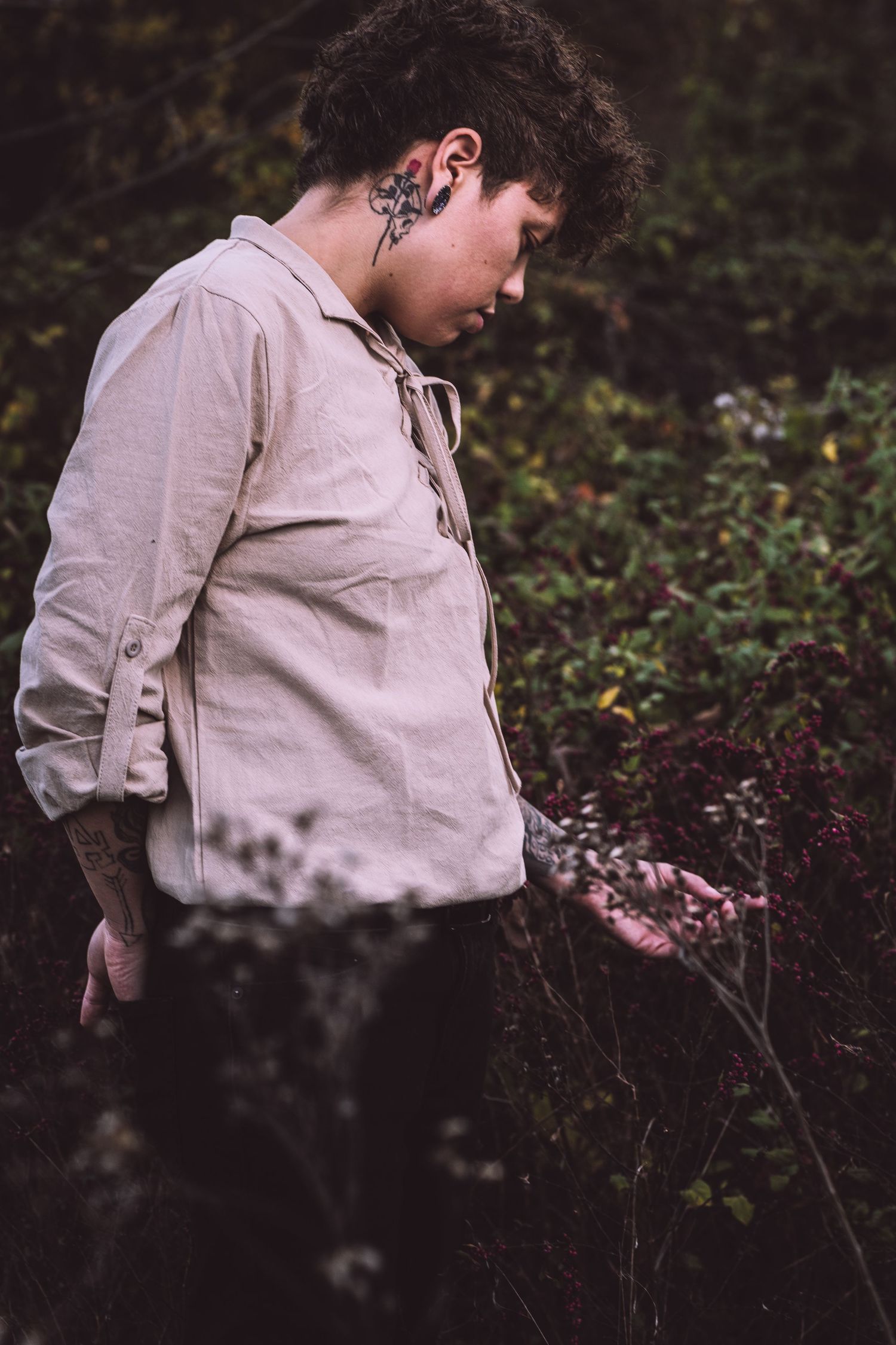 A figure in a light-colored shirt stands among foliage in a moody outdoor setting.