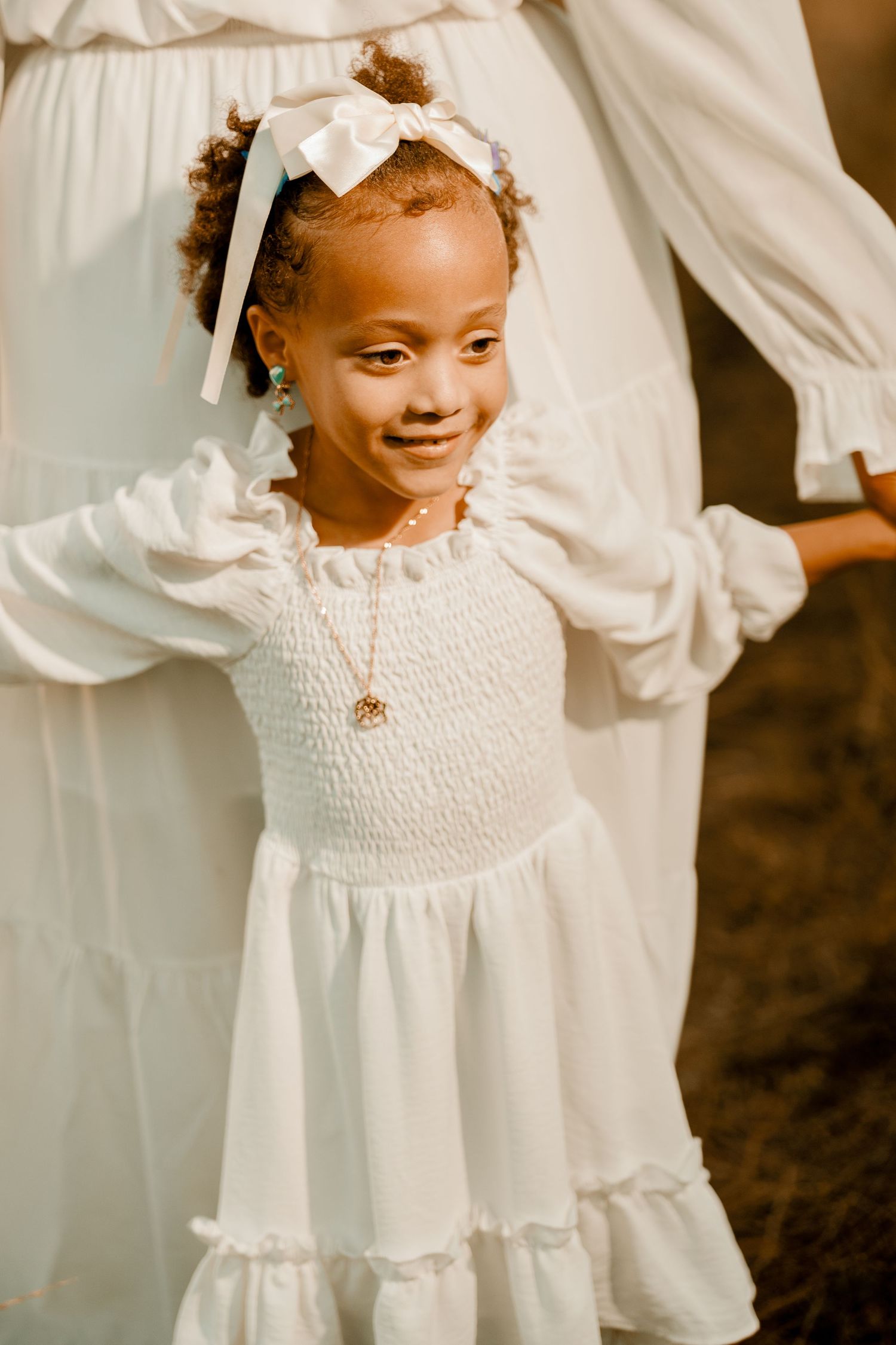 A young child in a white ruffled dress with a bow accessory smiles during an outdoor photoshoot.