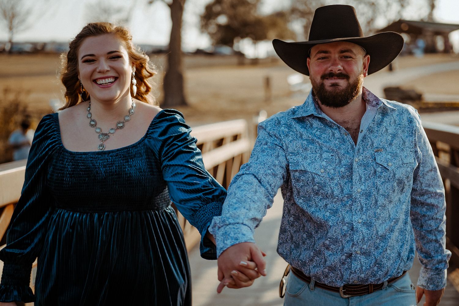 A couple in western attire, with her in a blue velvet dress and him in a patterned shirt and cowboy hat, share a joyful moment.