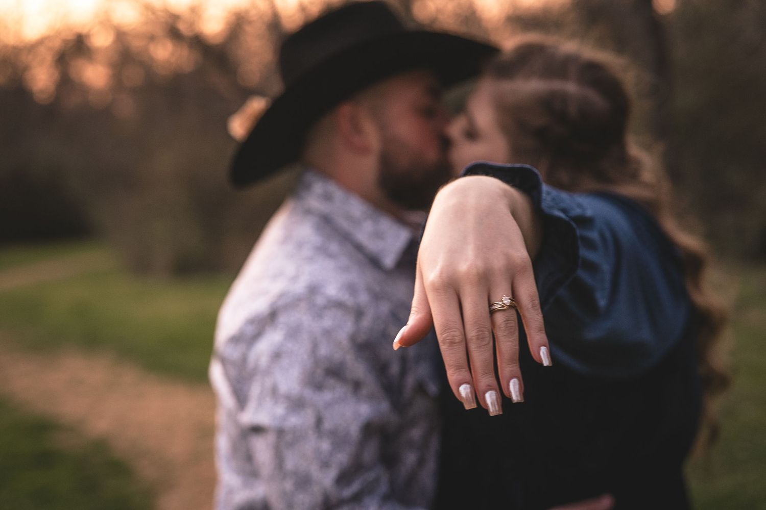 Person displaying engagement ring with partner in background during sunset.