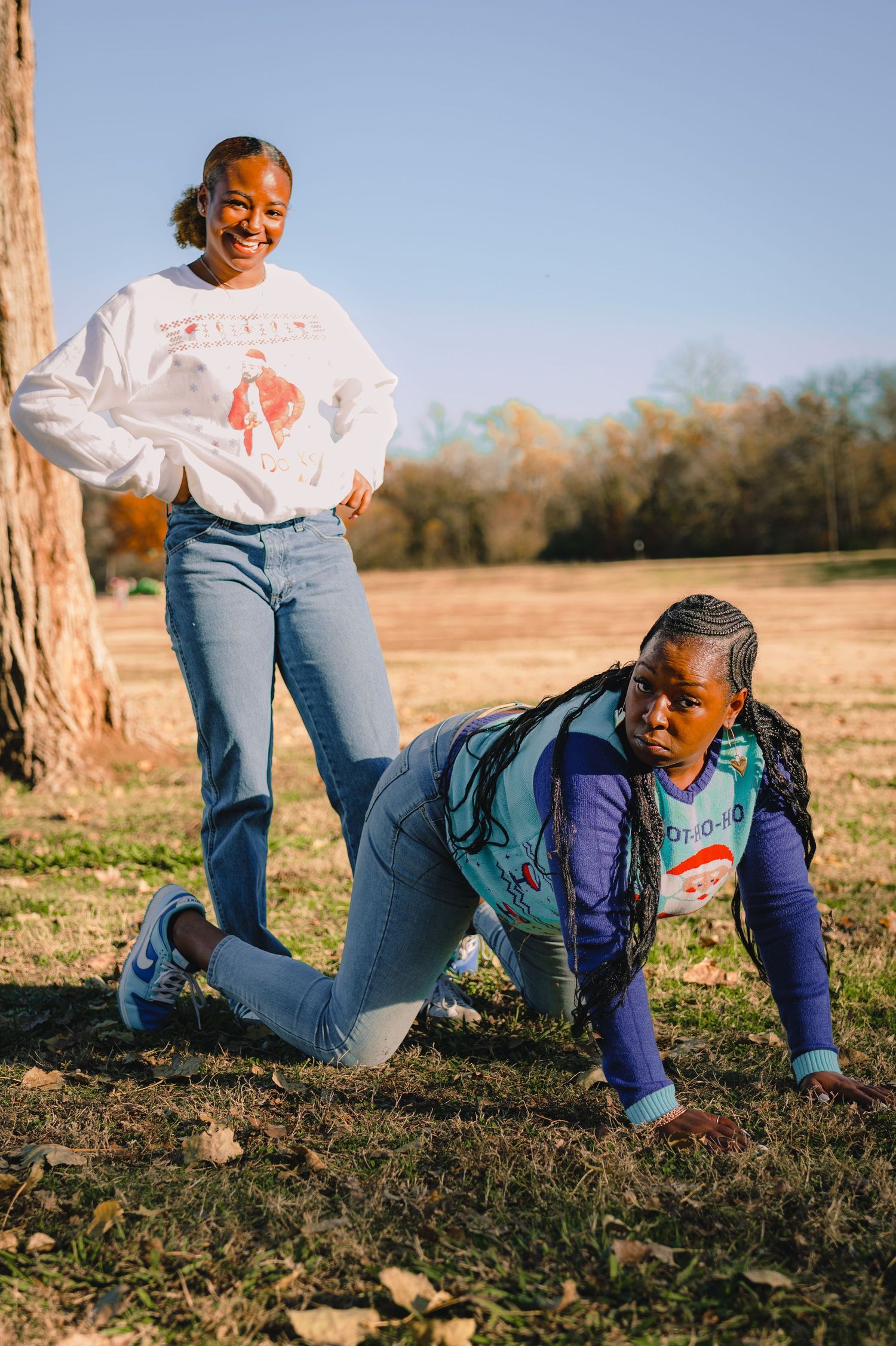 Two people in festive sweaters strike playful poses on grass near a tree.