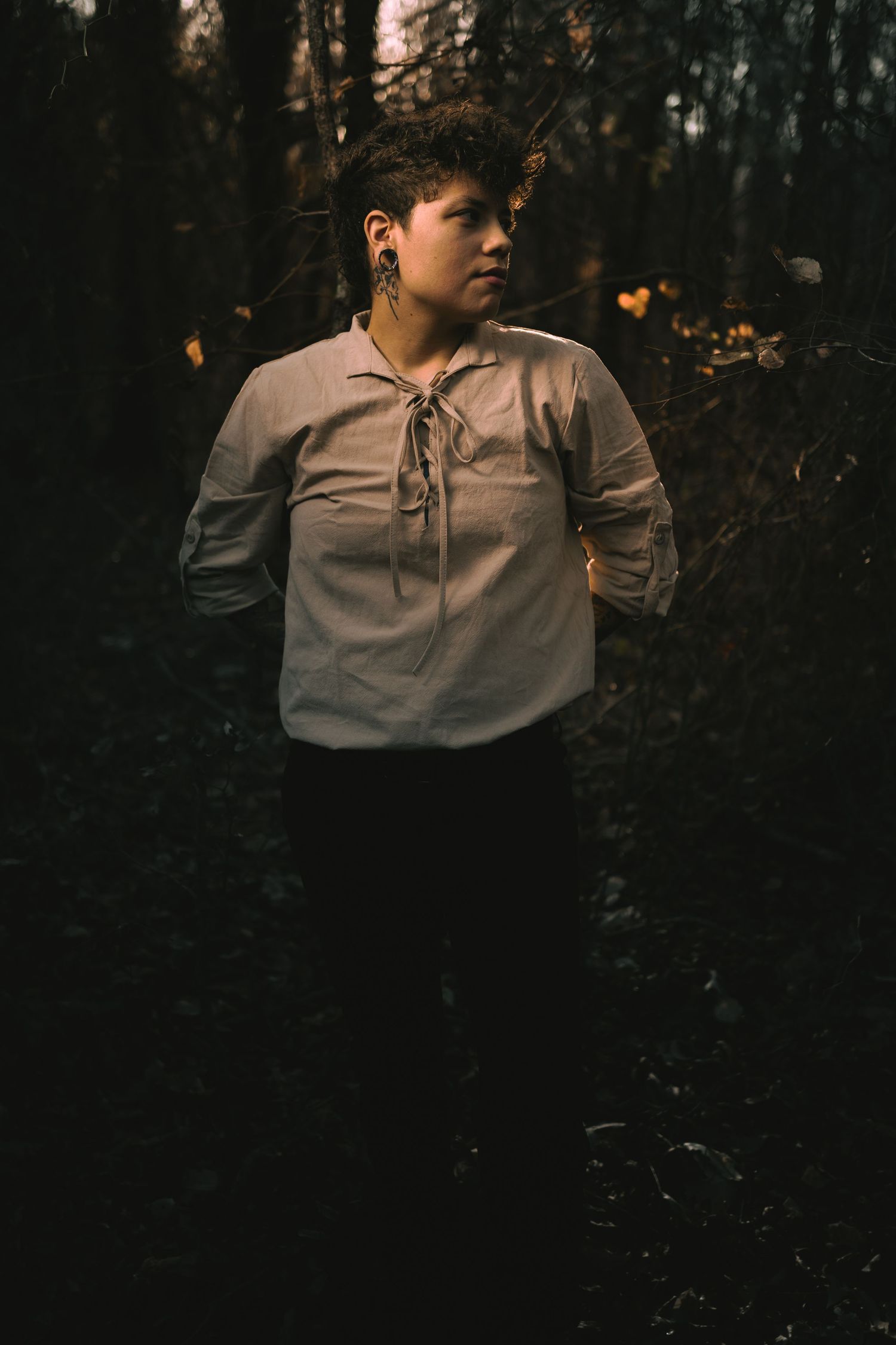 A figure in light clothing poses against a dark forest backdrop during sunset.