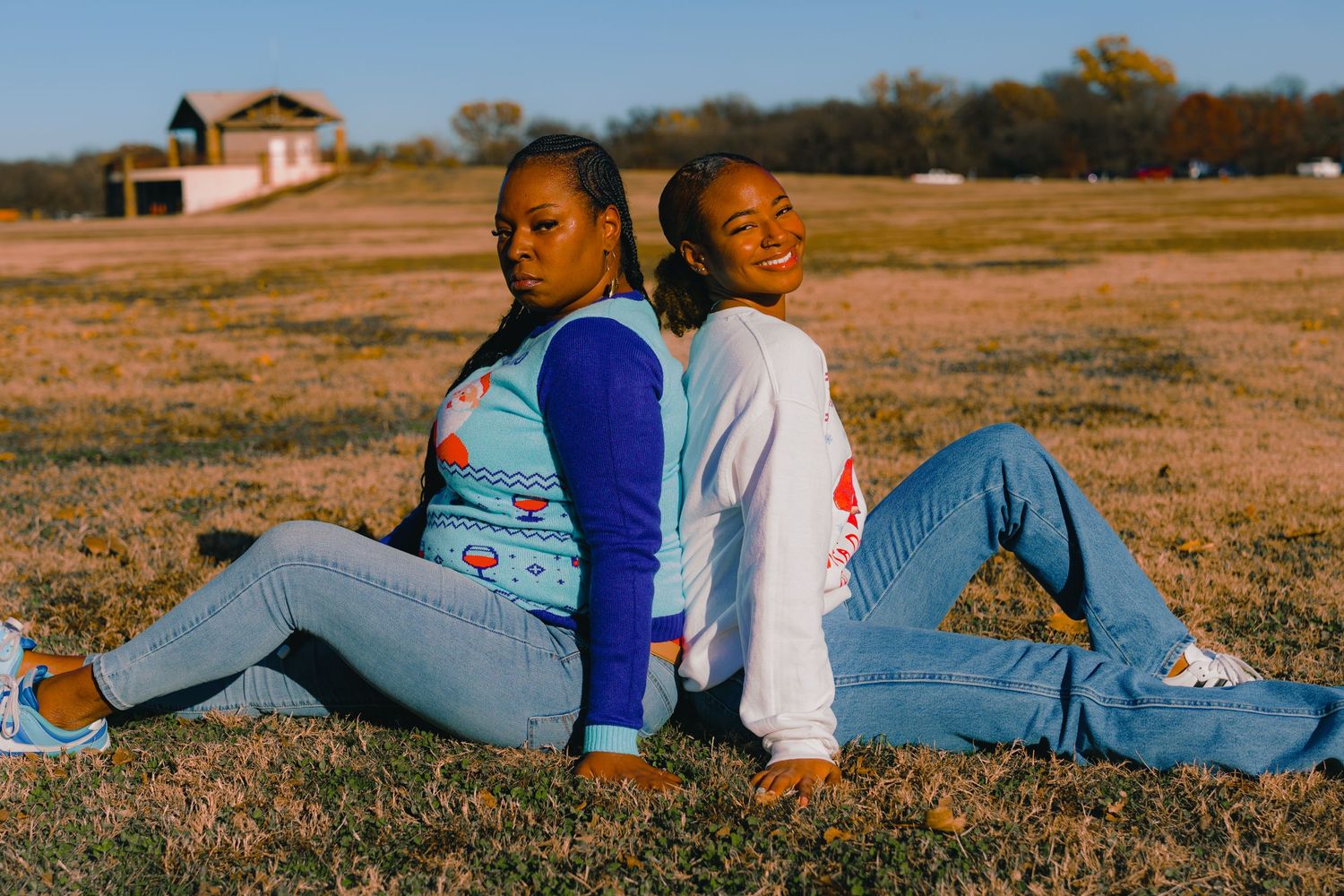 Two people wearing casual denim and blue outfits sit back-to-back on grassy field at sunset.