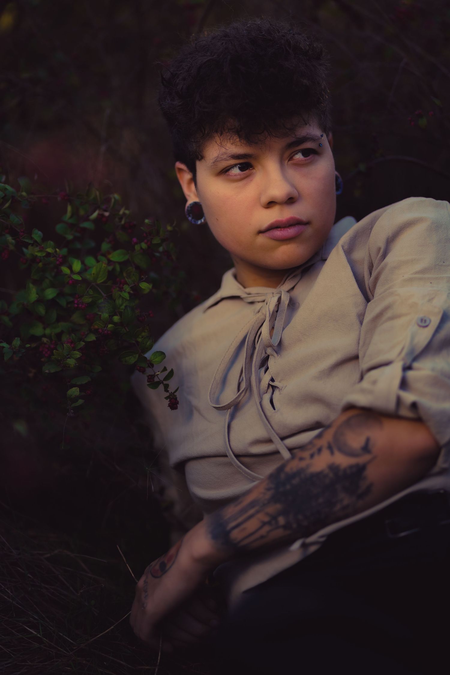 A pensive portrait showing tattoo art and vintage clothing against dark greenery.