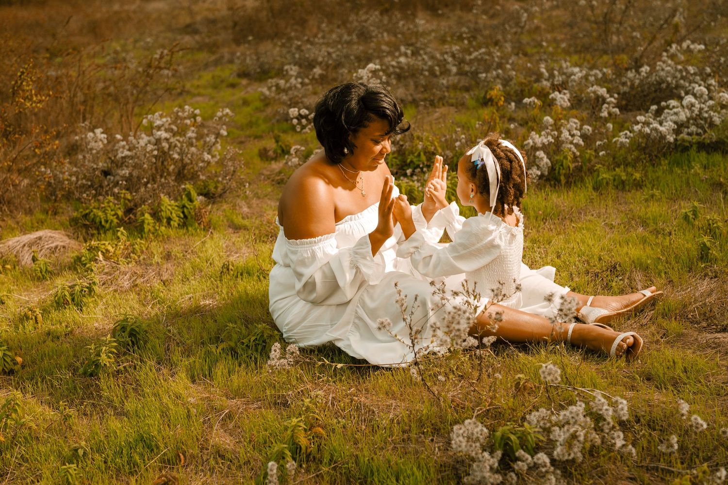 Two figures in ethereal white dresses share a tender moment among wildflowers at sunset.