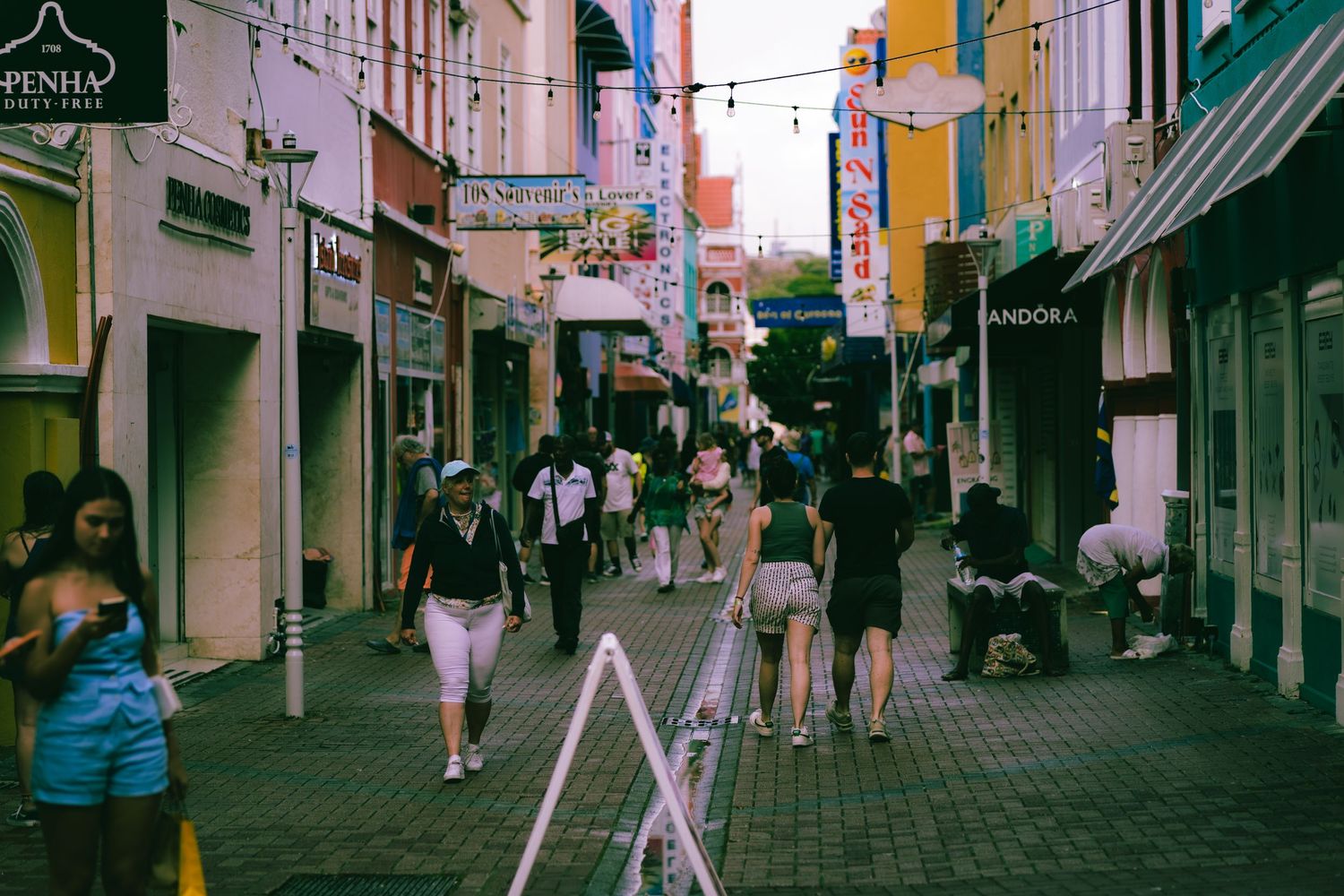 Bustling shopping street with colorful storefronts and pedestrians walking under string lights.