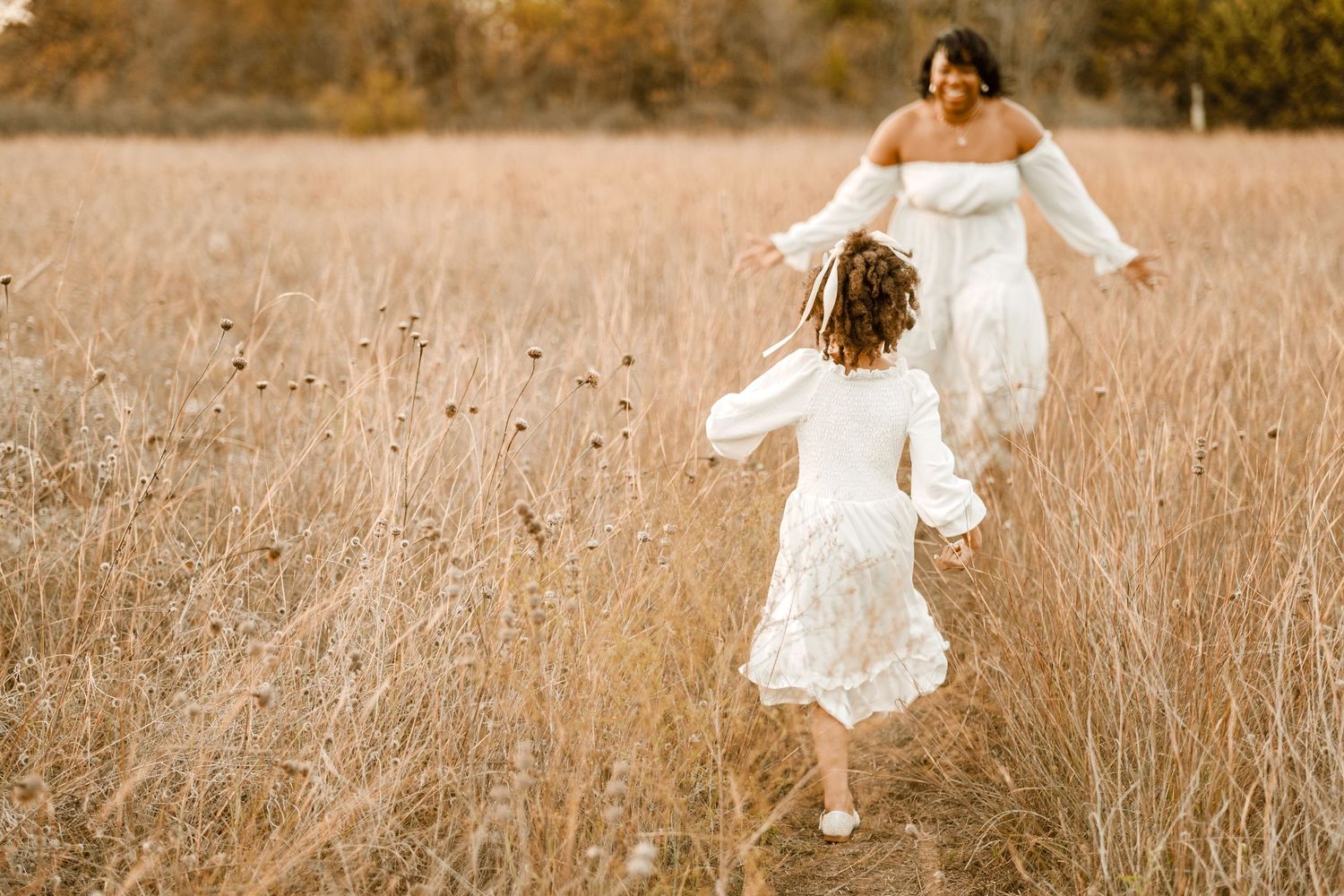 Two people in flowing white dresses run playfully through a golden autumn field.