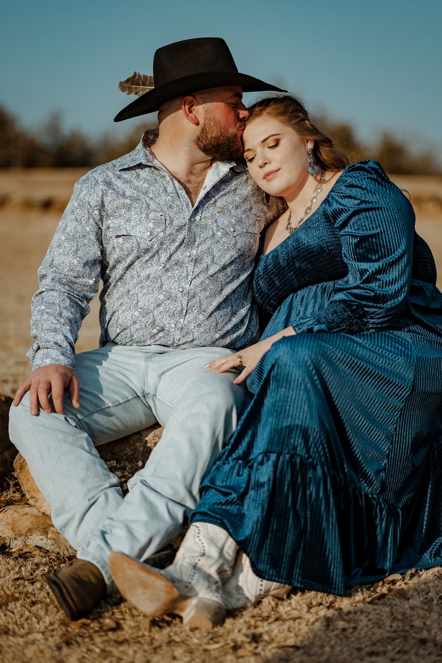 A couple in western attire share an intimate moment while sitting on rural ground.