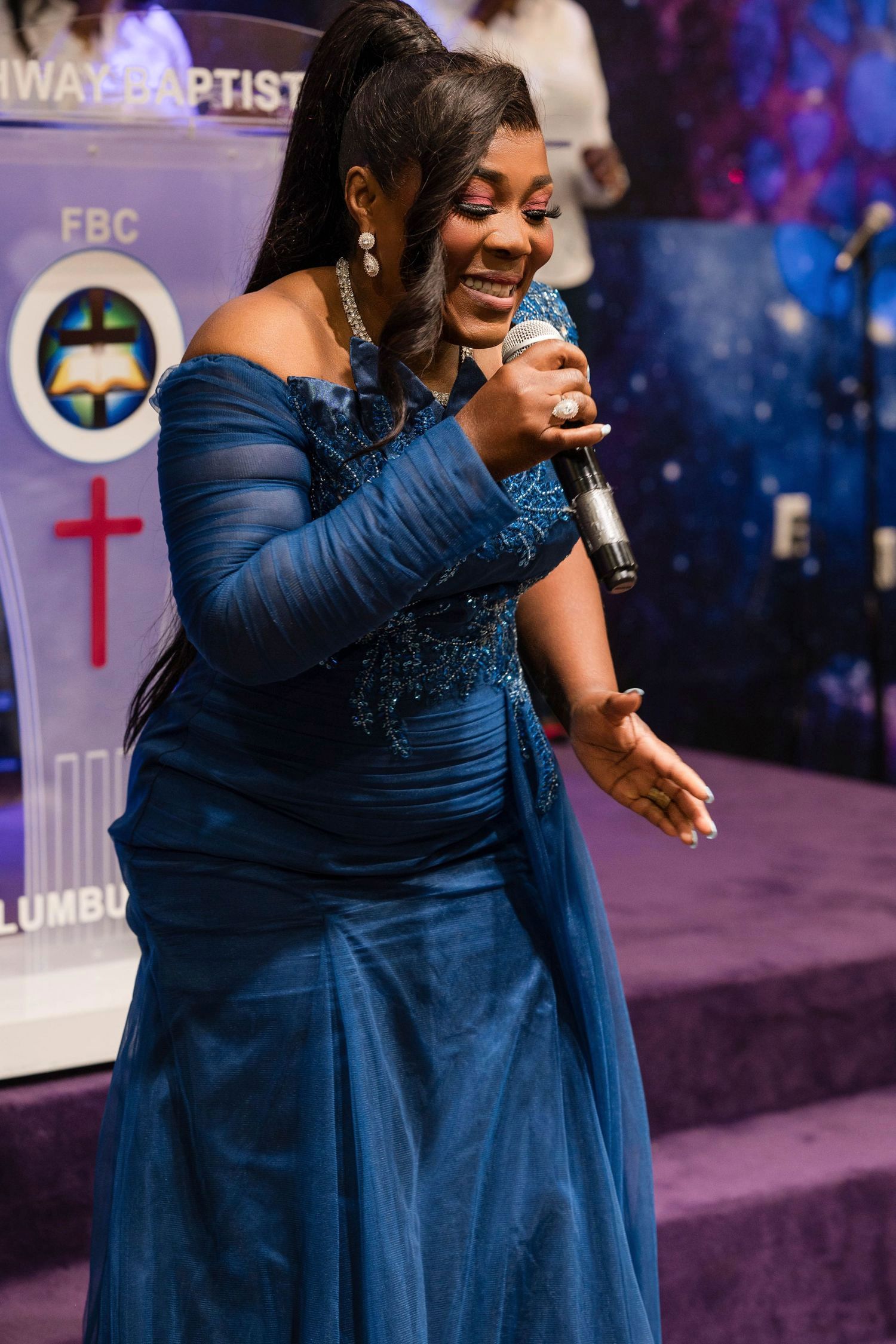 Singer in a navy blue evening gown performs passionately with a microphone on a purple-lit church stage.