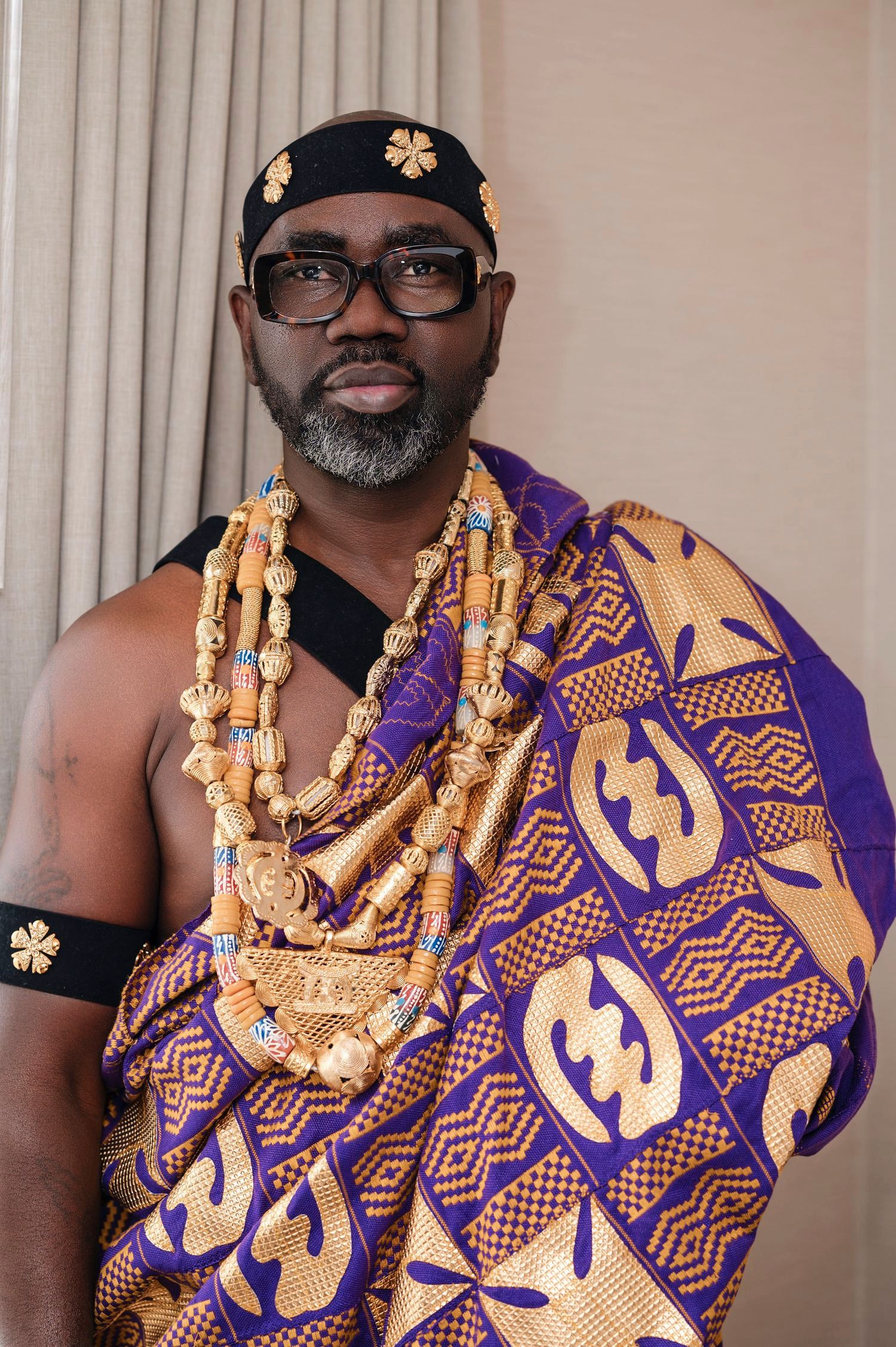 Person wearing traditional African attire with gold beaded necklaces and purple patterned kente cloth against light background.