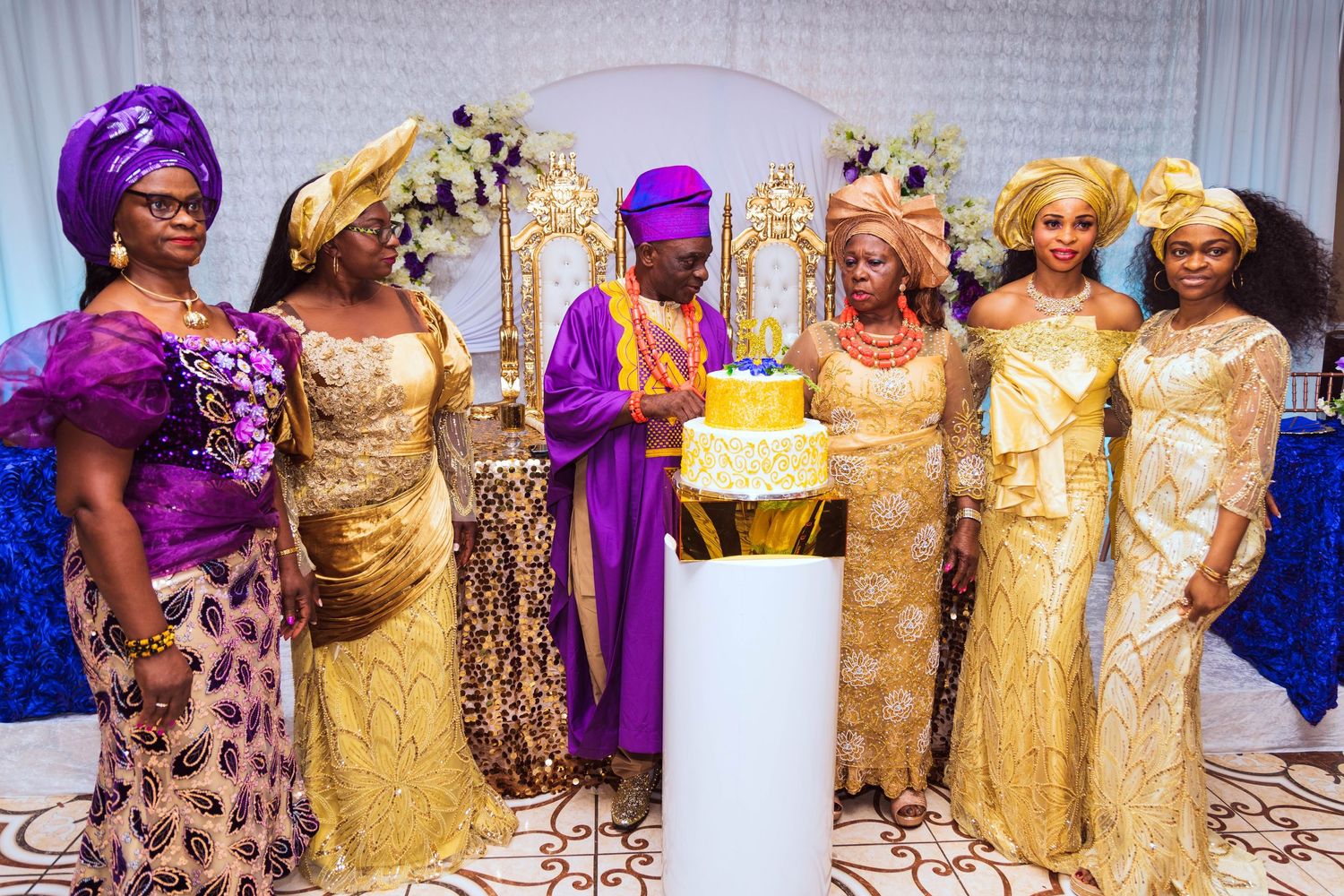 Women in vibrant gold and purple traditional dresses gather around a yellow cake at a formal celebration event.