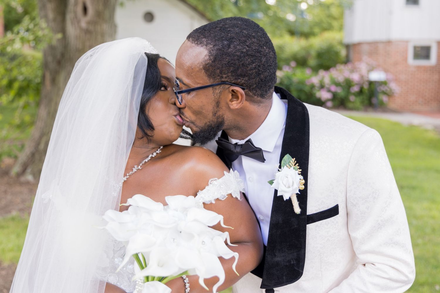 A romantic wedding portrait showing a tender moment between newlyweds in white attire by a garden.