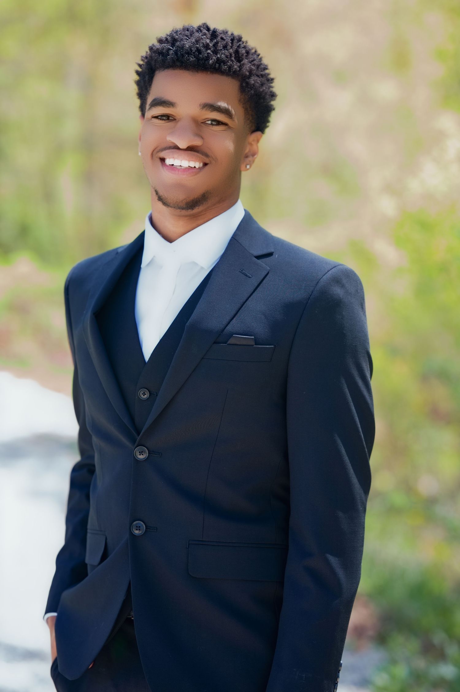 Happy portrait of a young man wearing a navy blue three-piece suit with white dress shirt in an outdoor setting.
