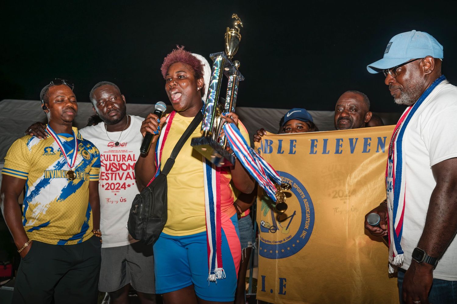 Athletes in yellow uniforms celebrate with large trophy during nighttime ceremony.