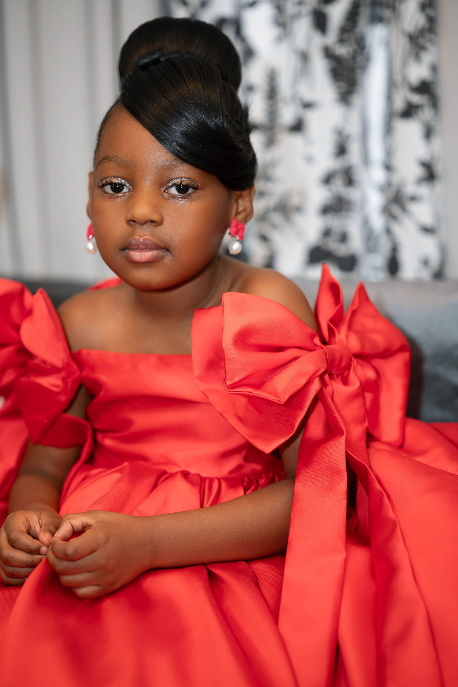 Child wearing a bright red formal dress with large bow details sits elegantly on a gray couch against floral curtains.