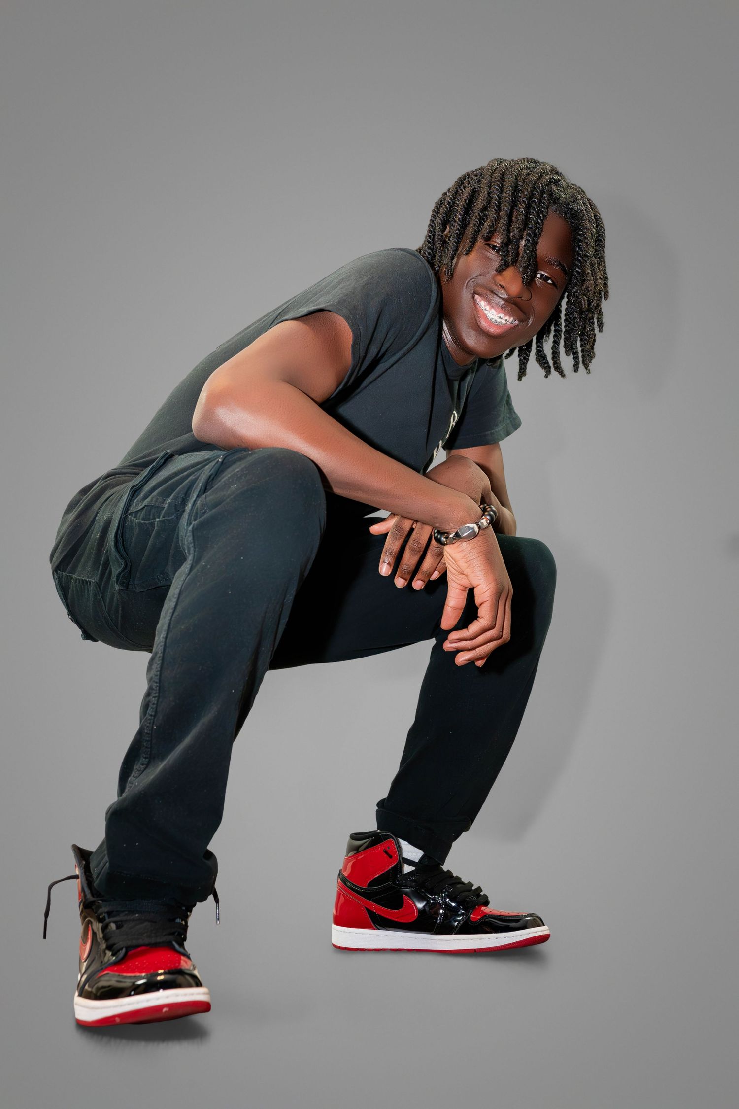 Dancer in dark athletic wear and red sneakers crouches with a bright smile against a grey studio backdrop.