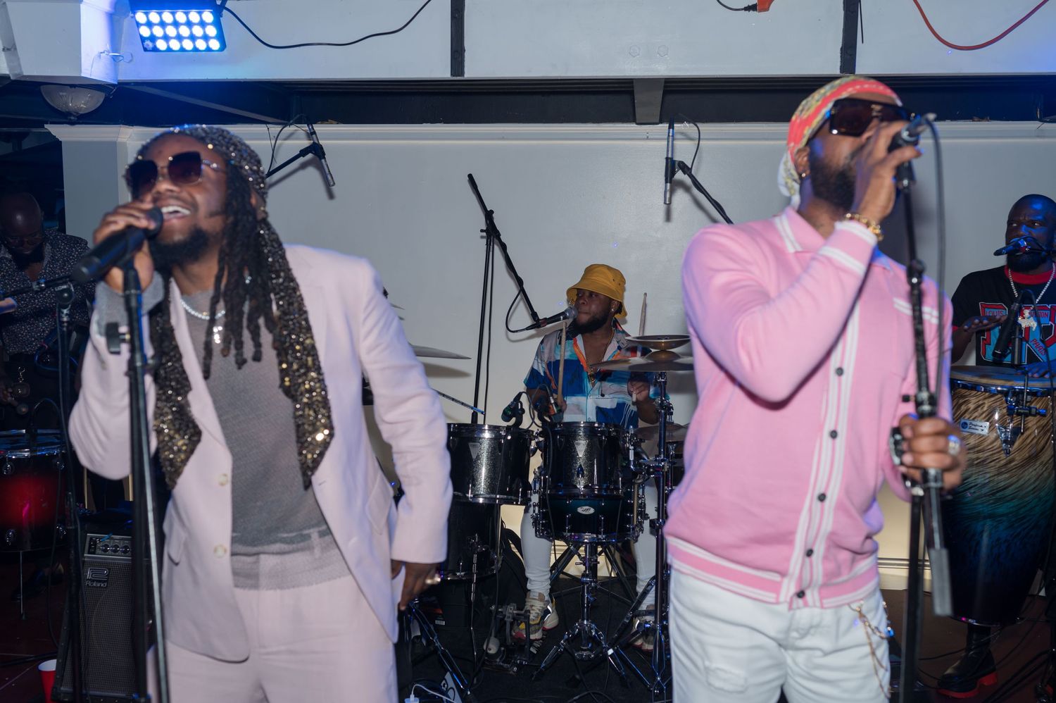 Two performers in light-colored outfits singing on stage with a band in the background.