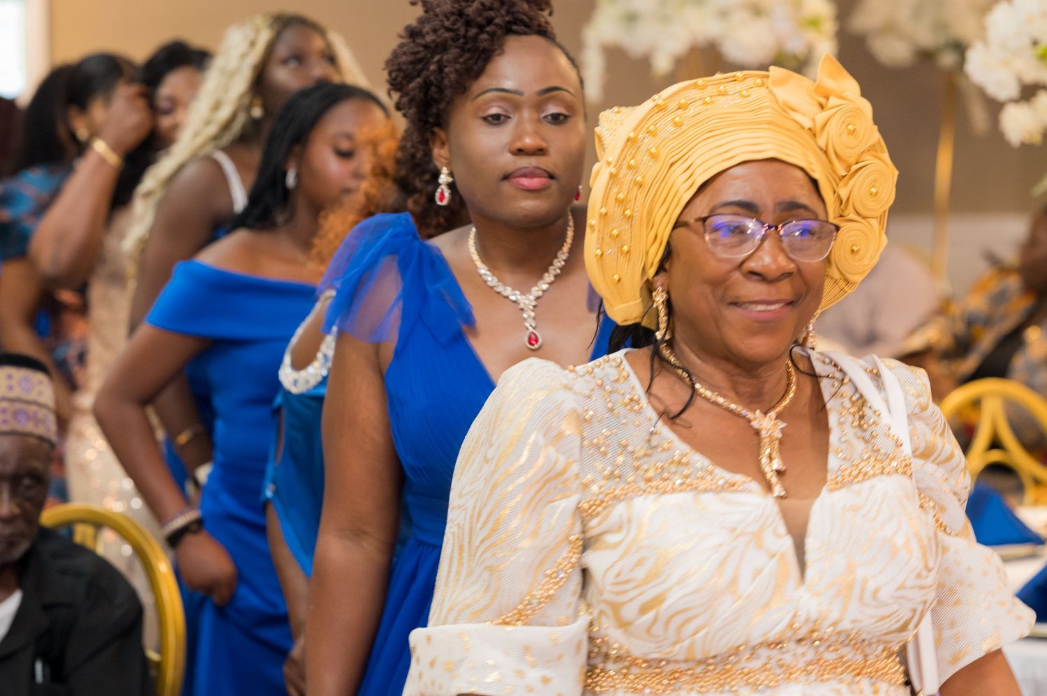 Guests in formal attire attending celebration with woman wearing yellow headpiece in focus.