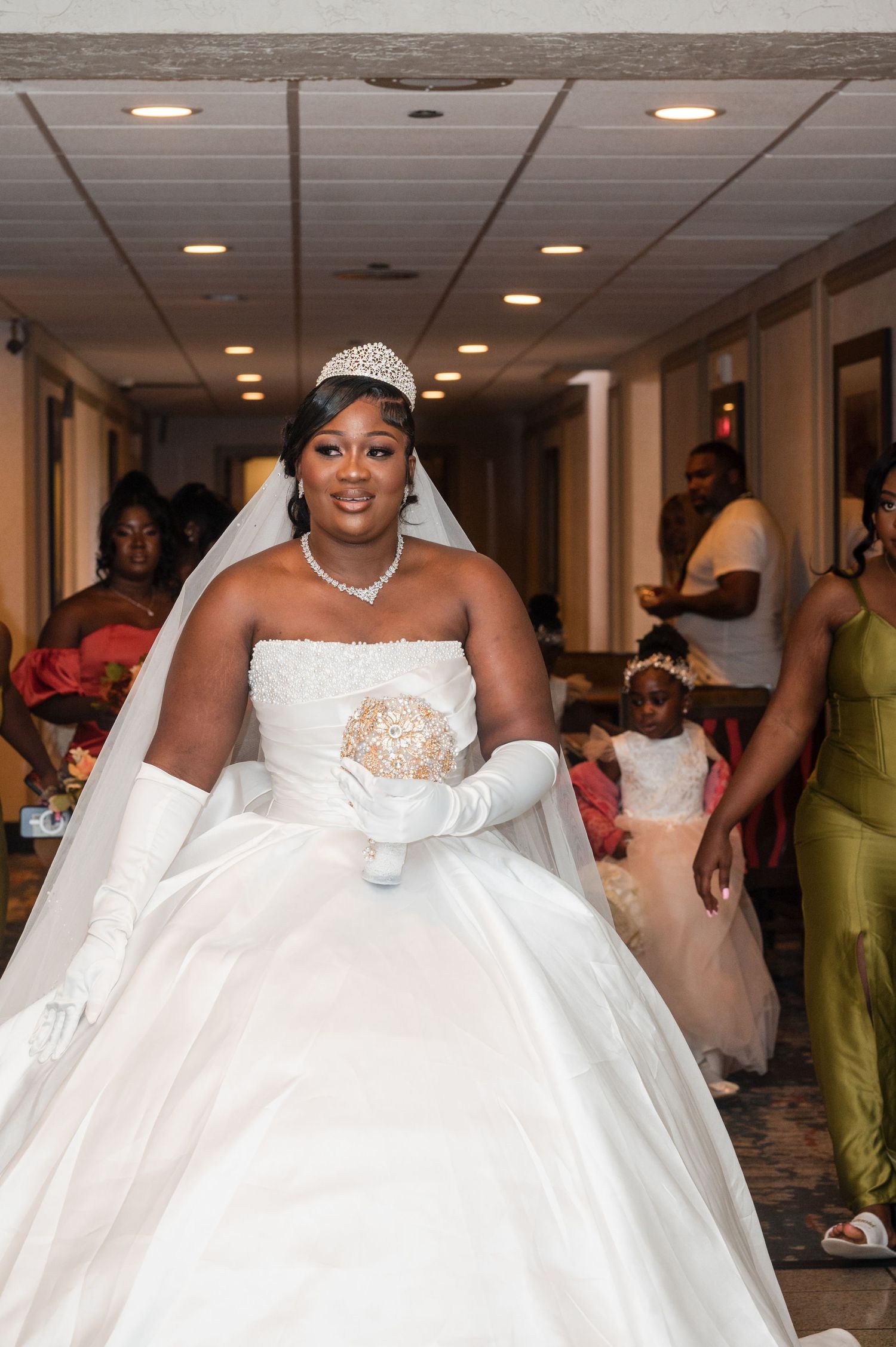 A bride in a glamorous ball gown with tiara walks down a hallway.