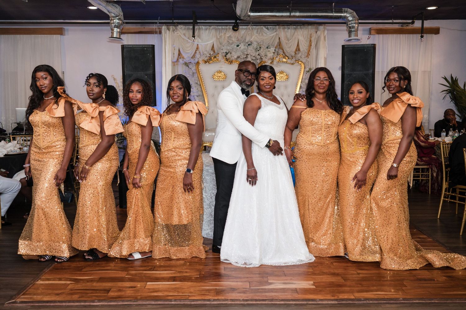 Wedding party photo with bridesmaids in matching gold dresses flanking couple at indoor reception.
