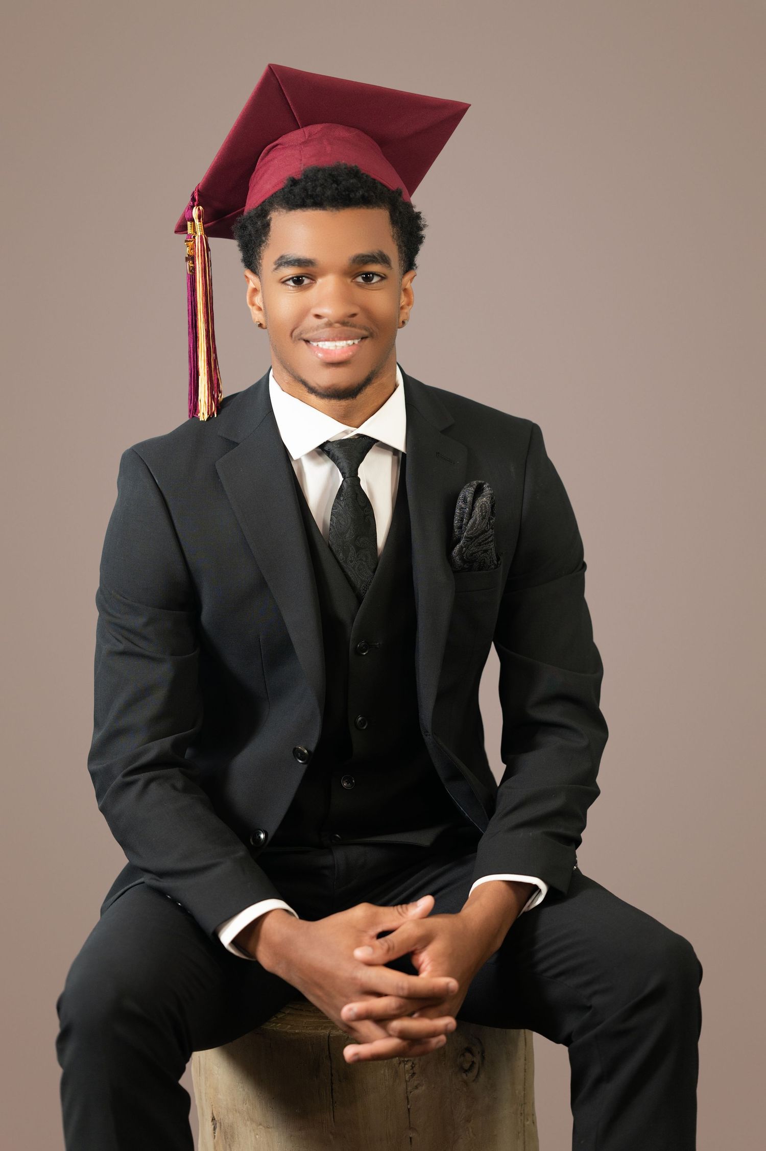 A graduate in formal black suit and burgundy cap sits on wooden block for professional portrait against neutral backdrop.
