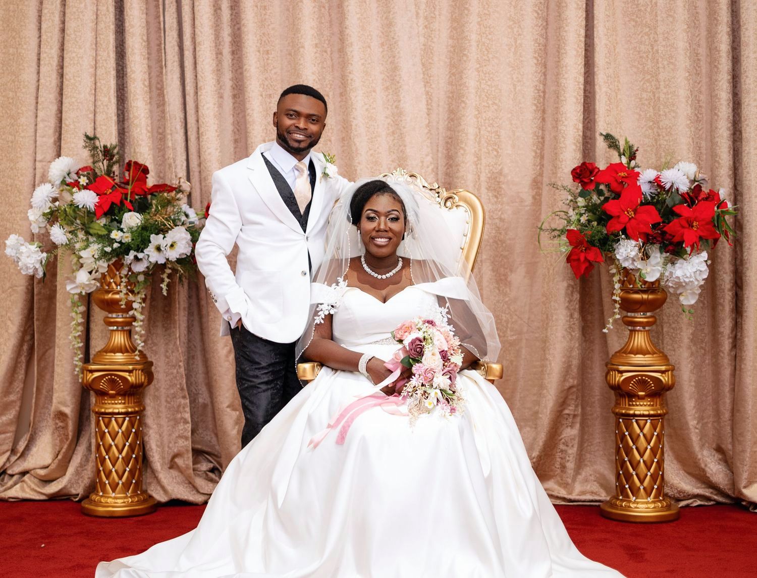 A wedding portrait with a couple in formal attire seated in front of beige curtains with red and white floral arrangements.