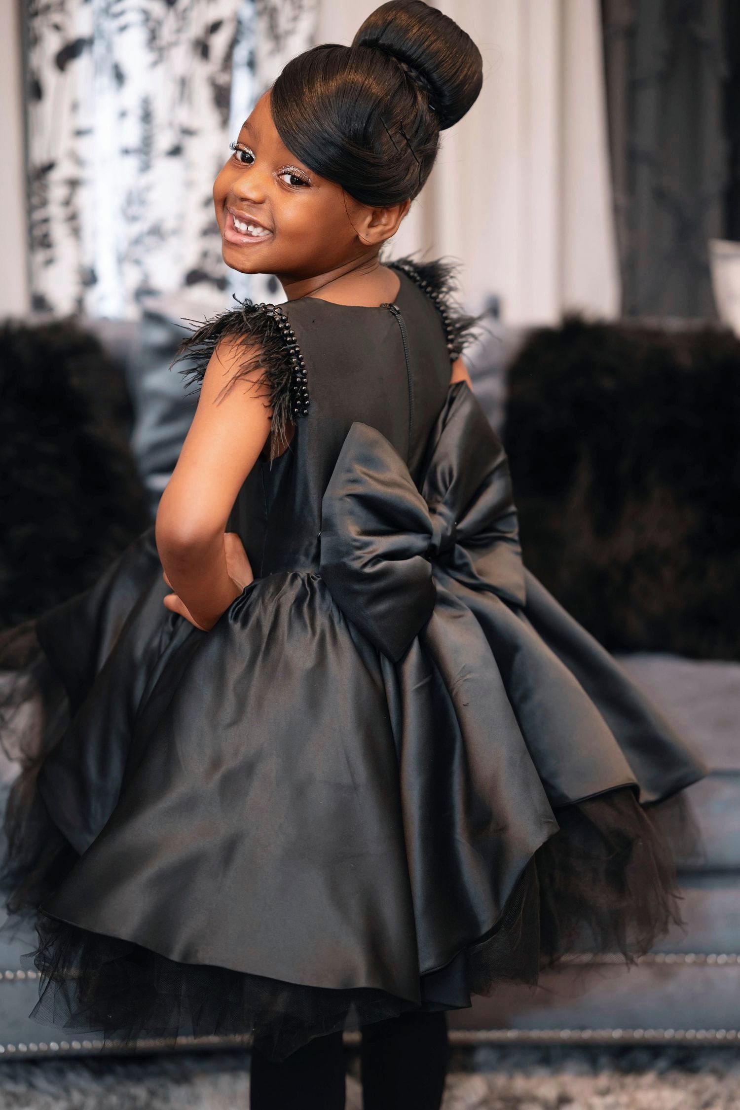 Elegant black party dress with tulle details and large bow accent shown from behind in indoor fashion photoshoot.