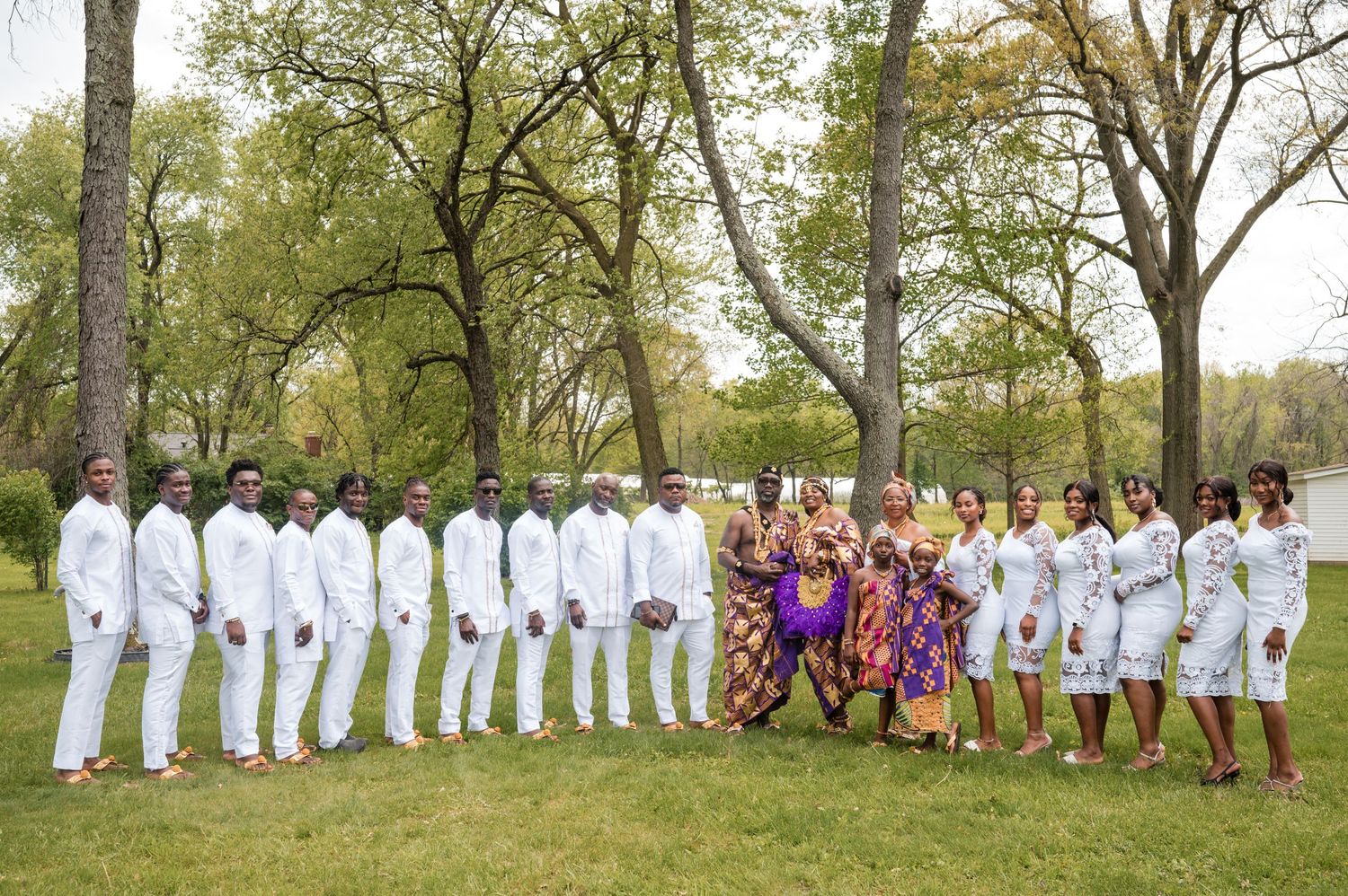Wedding party in white and purple attire poses outdoors among spring trees.
