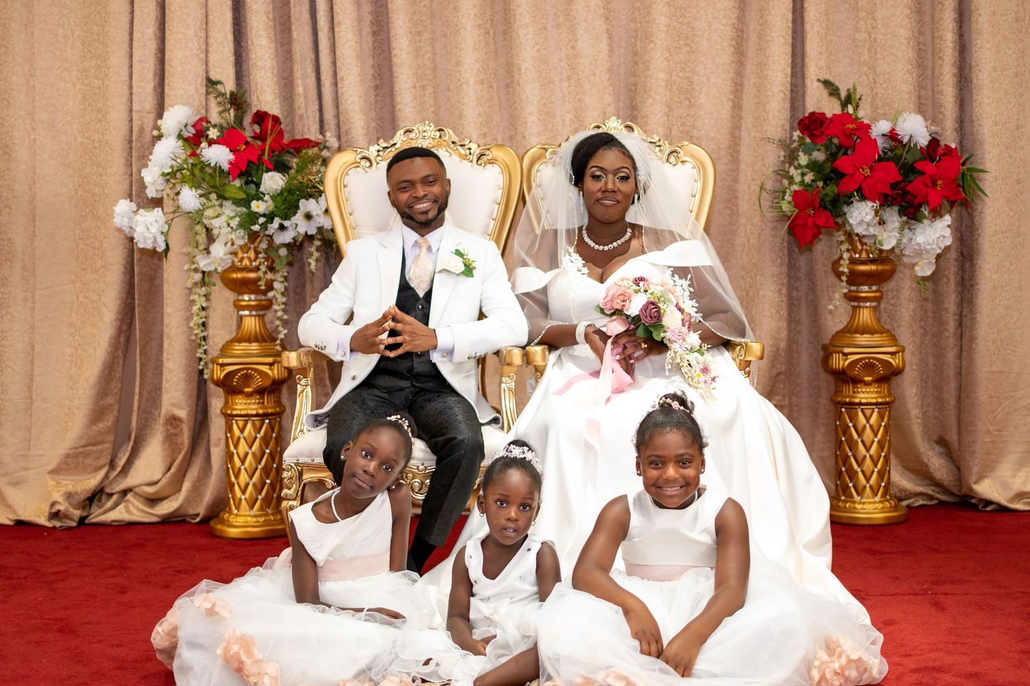 Elegant wedding group photo with ornate gold chairs, red and white floral arrangements, and rich burgundy backdrop.
