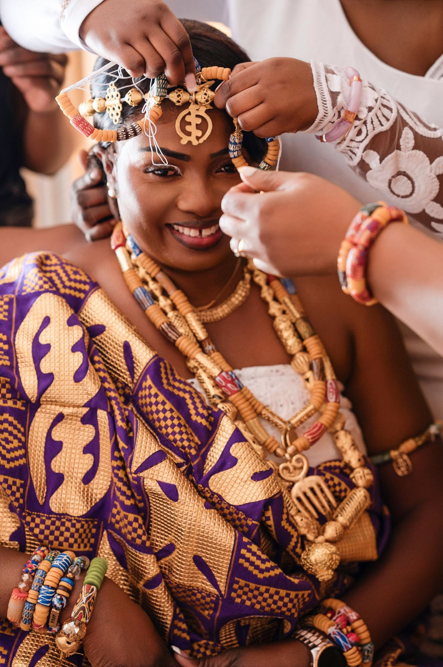 Traditional African bride wearing vibrant purple ankara dress adorned with gold jewelry and beaded accessories.