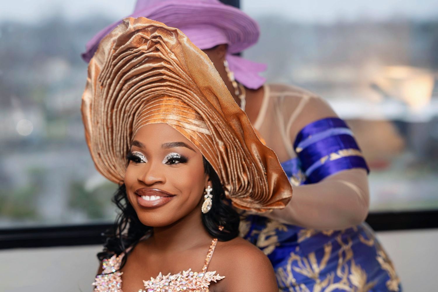 Woman in ornate gold headwrap and sparkly dress smiles while posing against blurred city backdrop.
