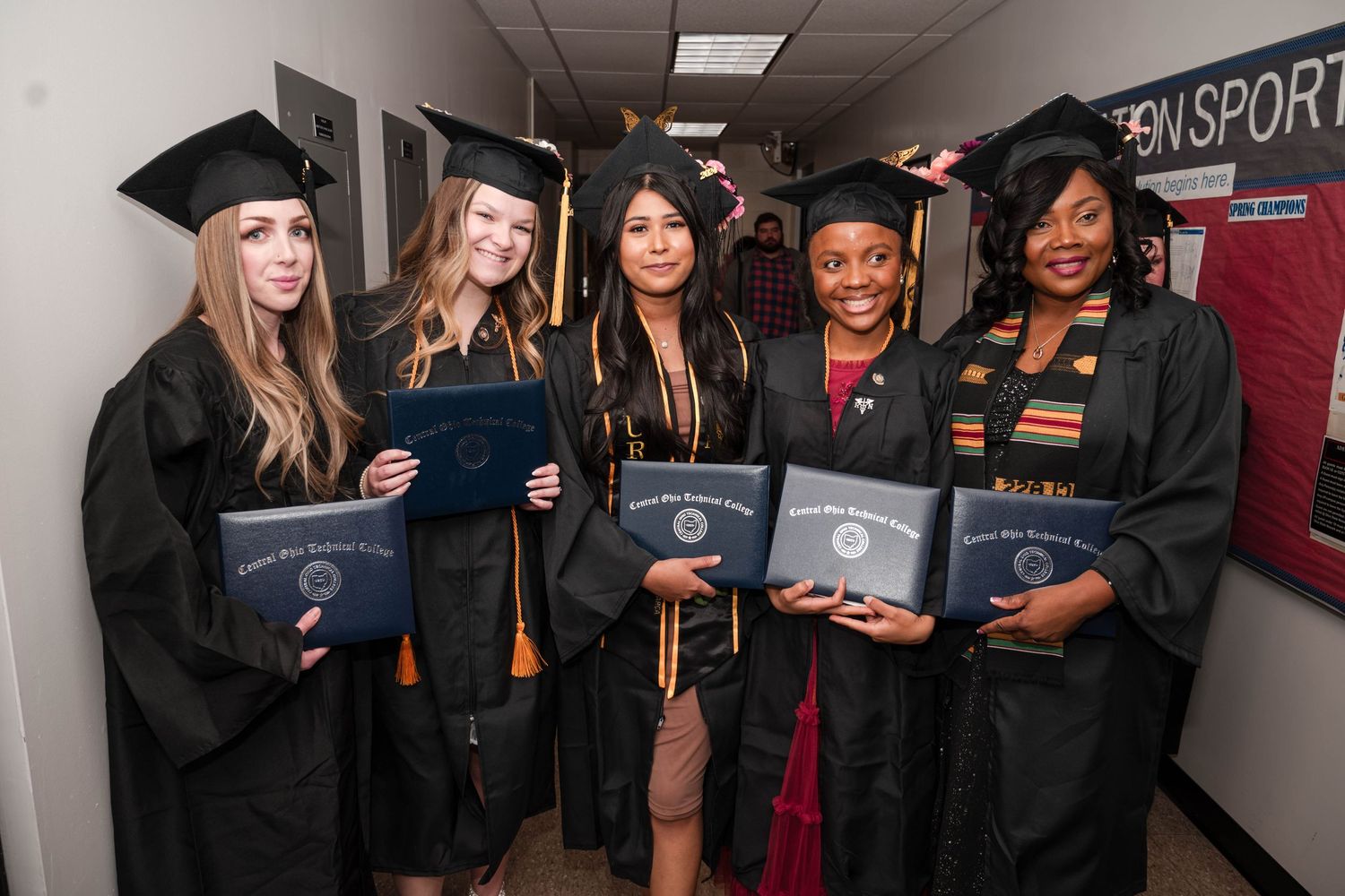Five graduates display their diplomas while wearing academic regalia in a hallway.