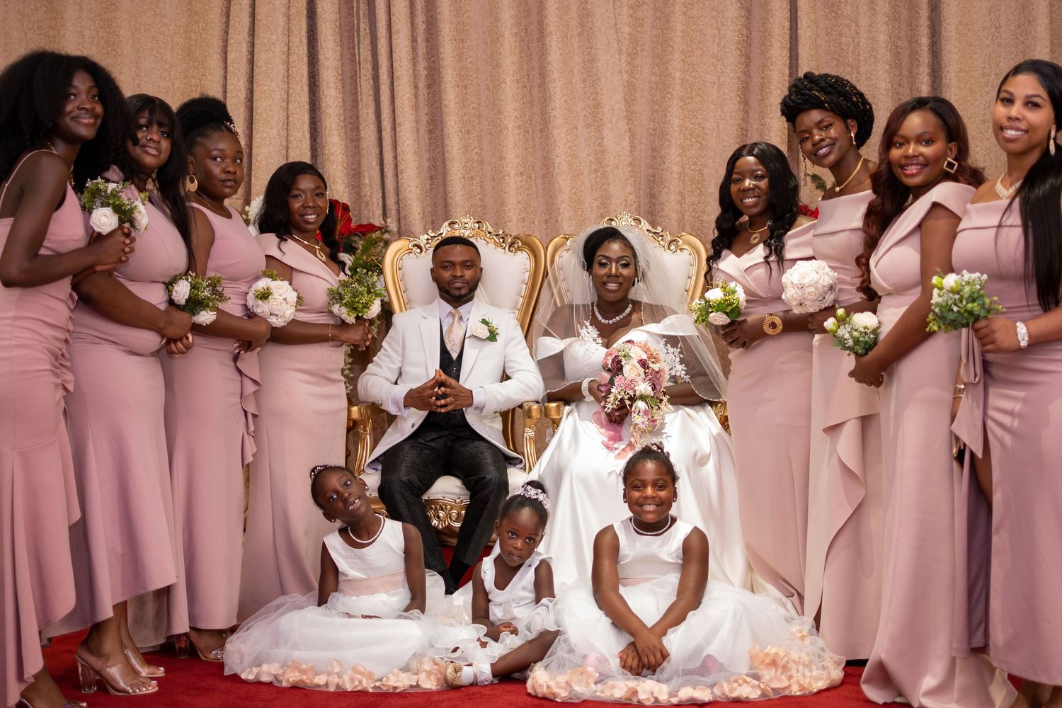 Wedding party in matching blush pink dresses poses together for a formal portrait on red carpet.