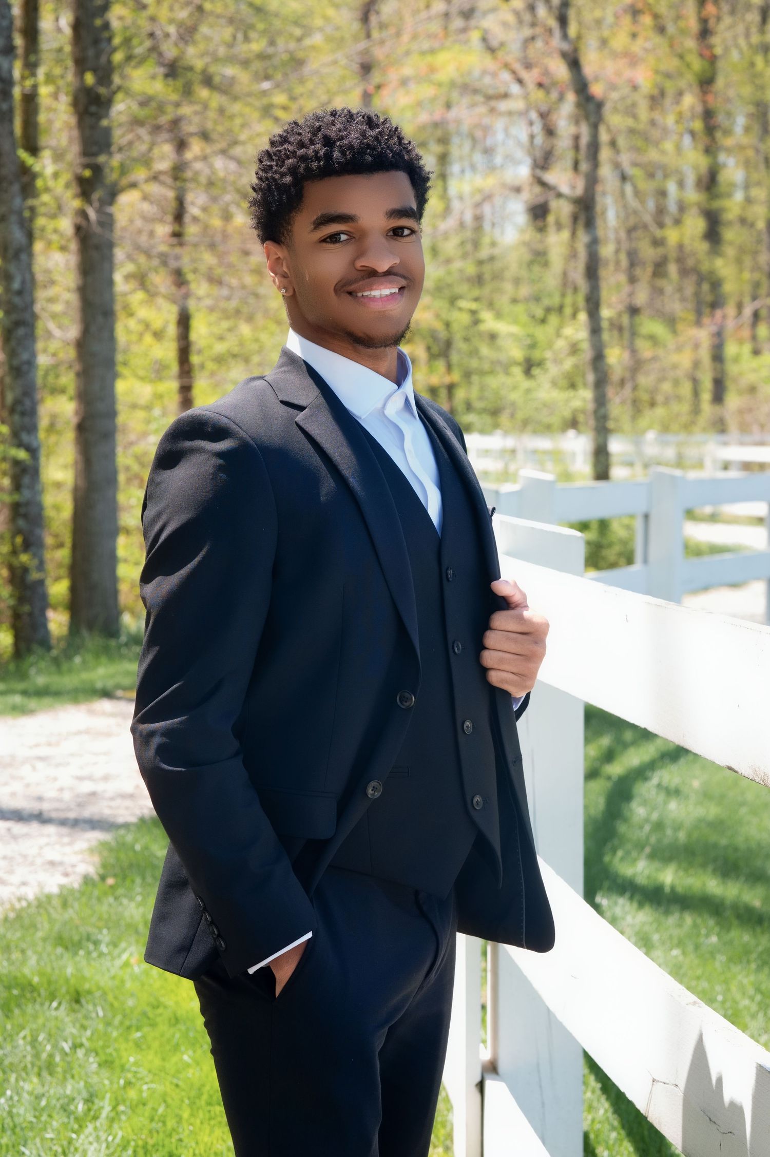 Young man poses outdoors in a navy suit against white fence and spring woodland background.