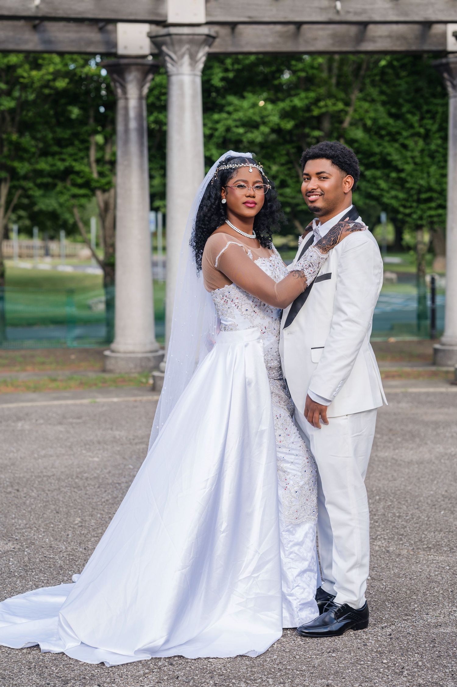 A couple in elegant white wedding attire pose together beneath classical stone columns in an outdoor setting.