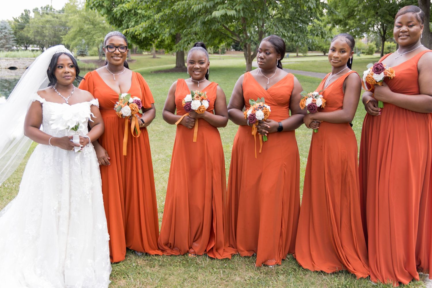 Bride in white gown stands with bridesmaids wearing matching orange dresses outdoors.