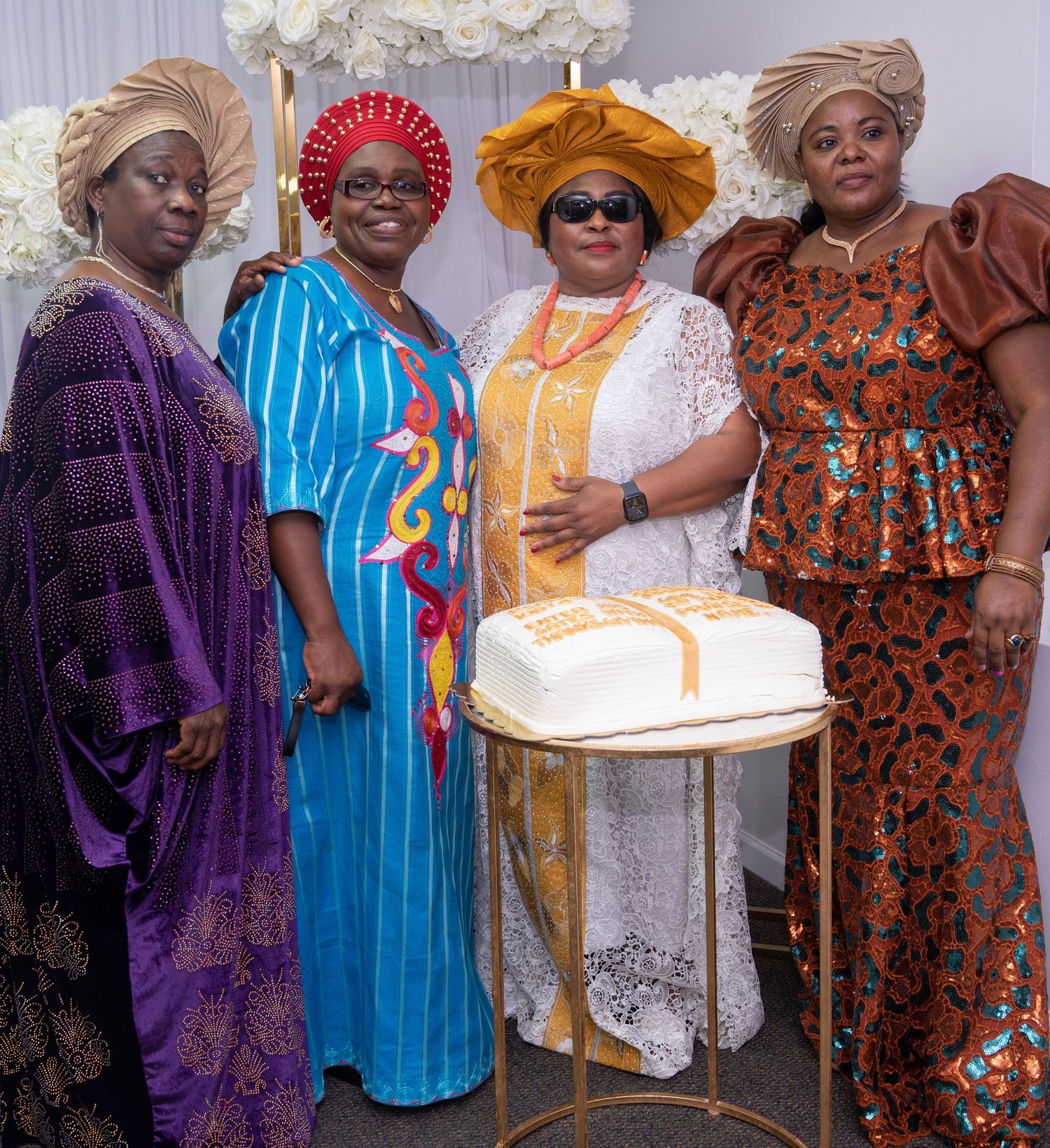 Four women in vibrant formal African attire stand beside a white cake at a celebration.