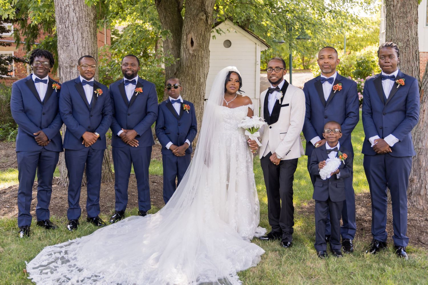 Wedding party in navy suits and long white dress pose outdoors under trees.