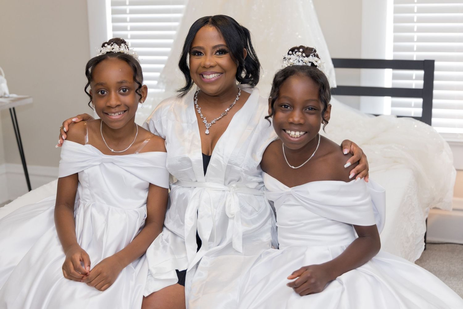 Three people in white formal attire pose together wearing tiaras.