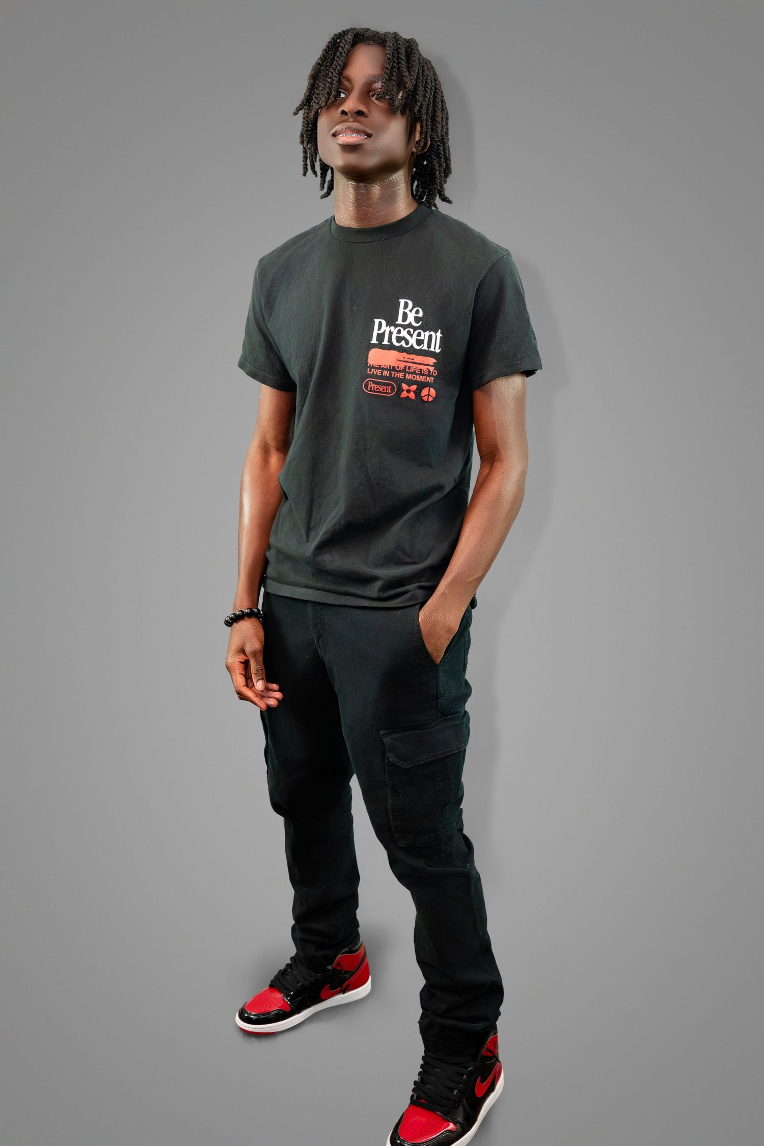 A person in a dark t-shirt and cargo pants poses against a gray backdrop wearing red and black sneakers.