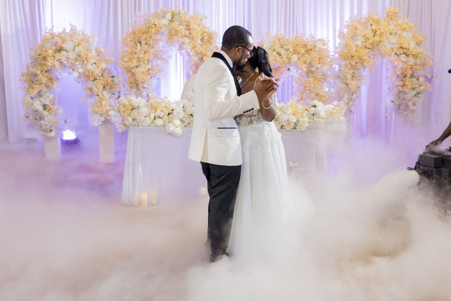 A couple share their first dance surrounded by white floral arches and romantic fog on their wedding day.